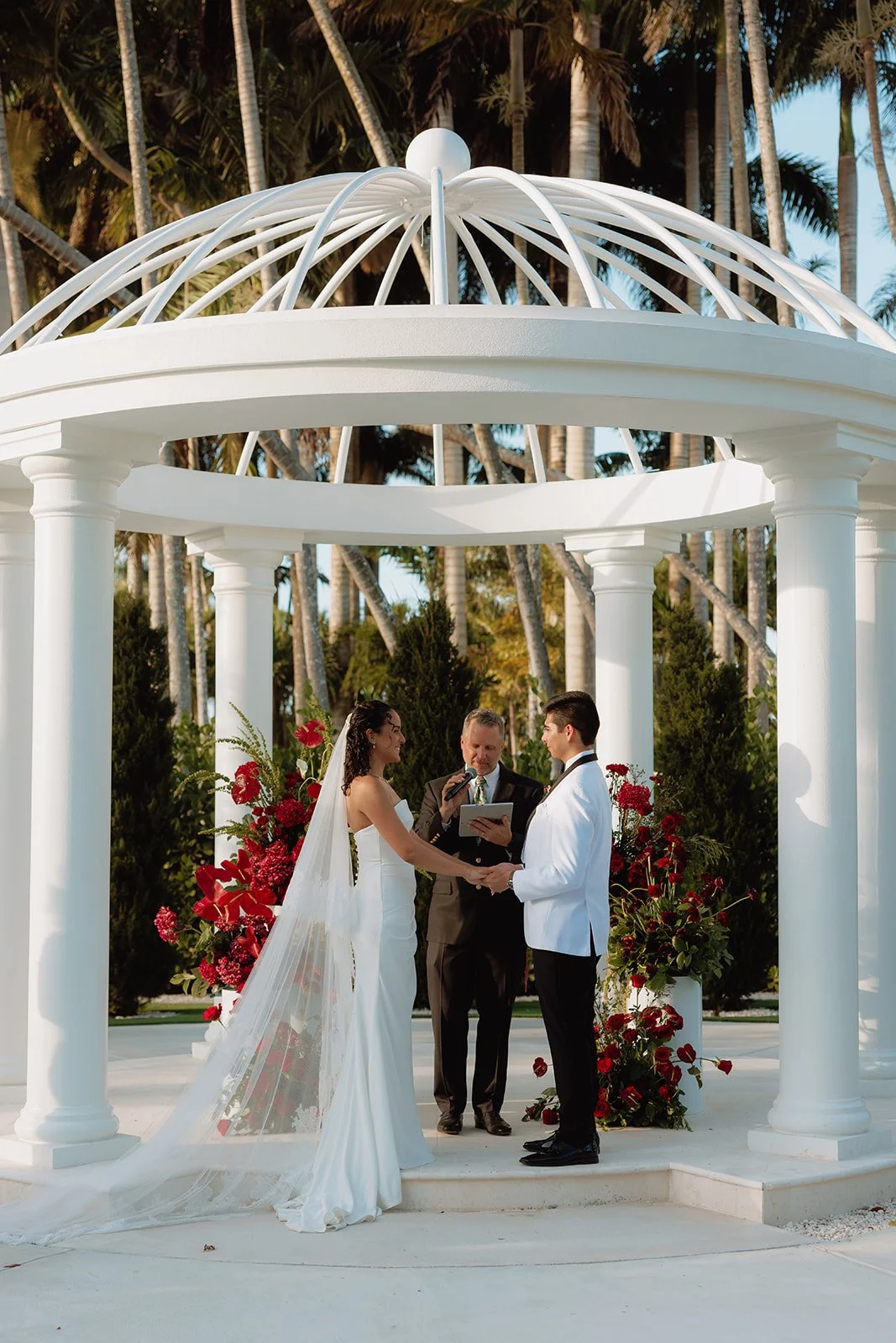 Bride and groom sharing a kiss during sunset while holding a cascading orchid and red rose bridal bouquet designed by Atico Floral, a luxury South Florida wedding florist.