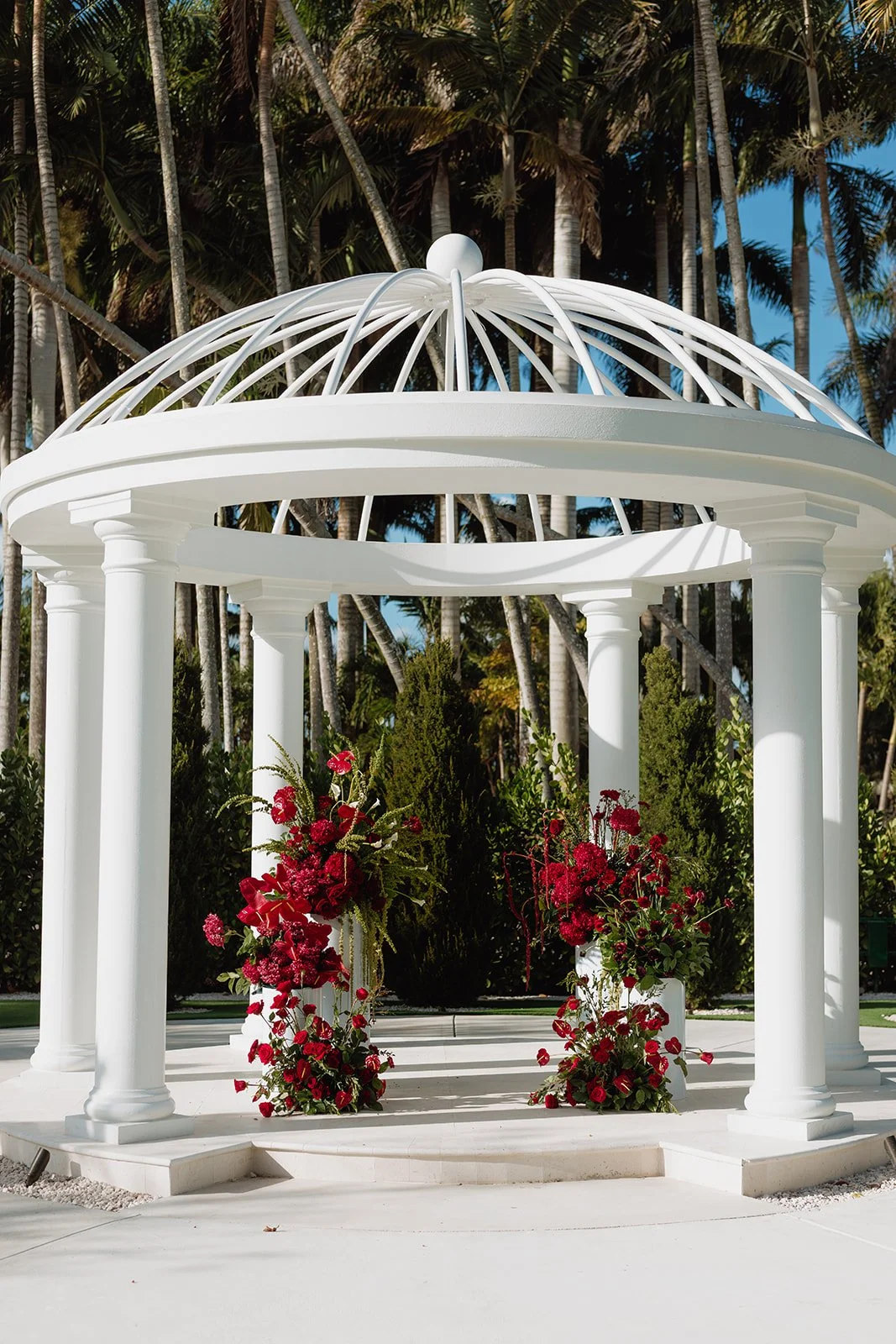 Luxury outdoor wedding ceremony featuring elegant red floral pedestal arrangements beneath a white gazebo, designed by Atico Floral, a Miami and South Florida wedding florist.