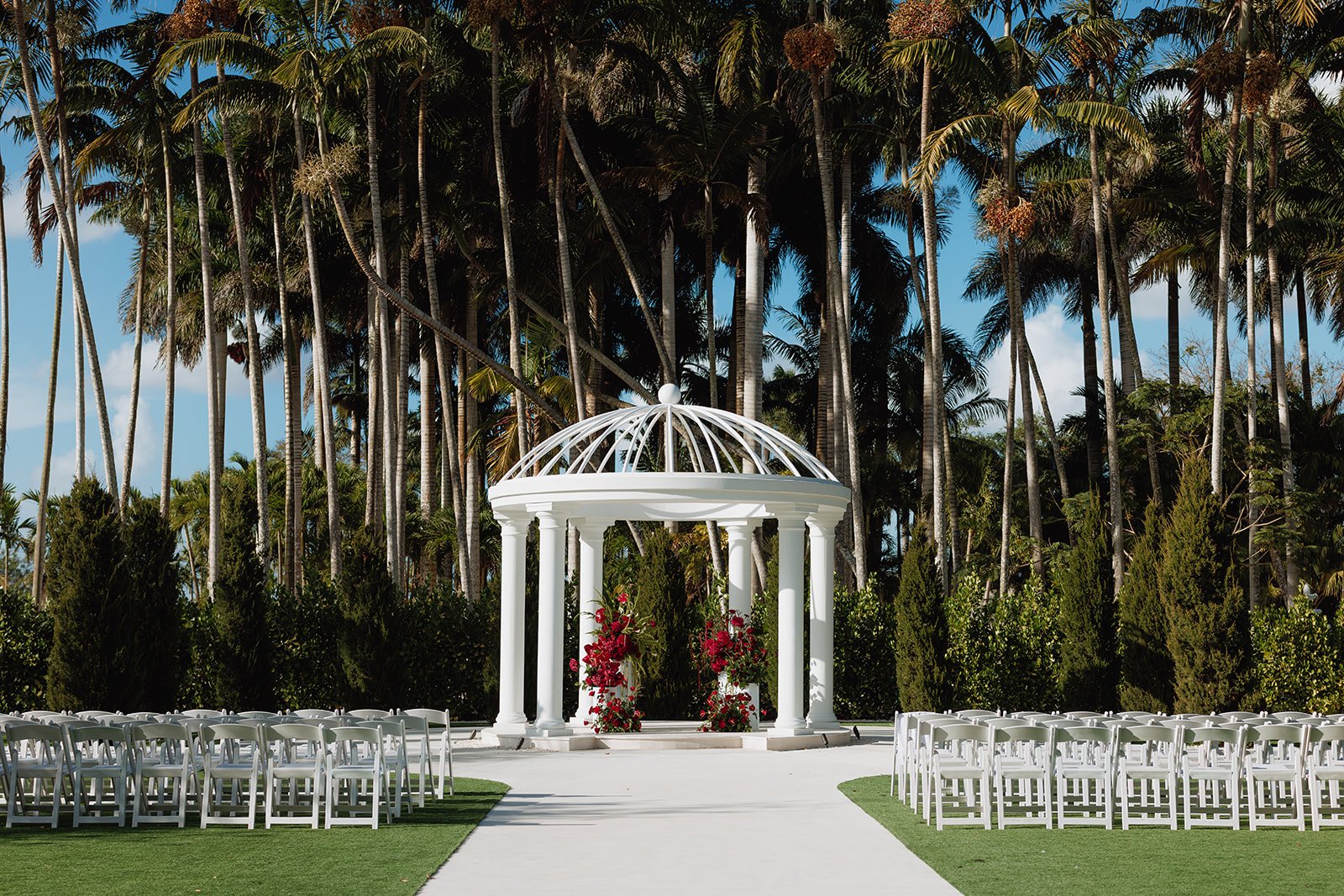 Luxury outdoor wedding ceremony featuring elegant red floral pedestal arrangements beneath a white gazebo, designed by Atico Floral, a Miami and South Florida wedding florist.