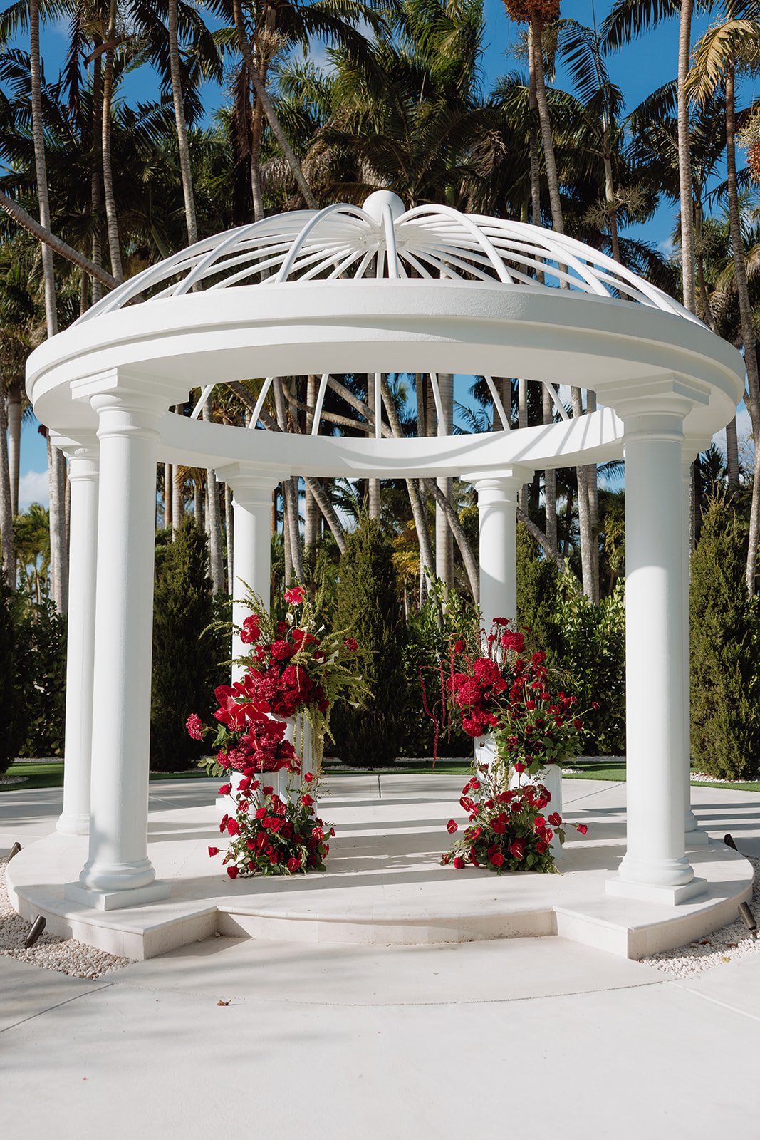 Bold, romantic ceremony florals designed beneath a classic white gazebo, featuring lush red roses, orchids, and cascading blooms. Atico Floral creates luxury wedding ceremony designs throughout Miami, Palm Beach, and South Florida, transforming beaut