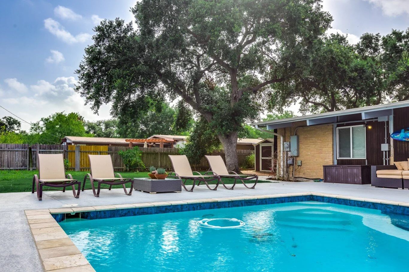 Backyard scene with a swimming pool, four lounge chairs, a tree, and a house with outdoor seating.