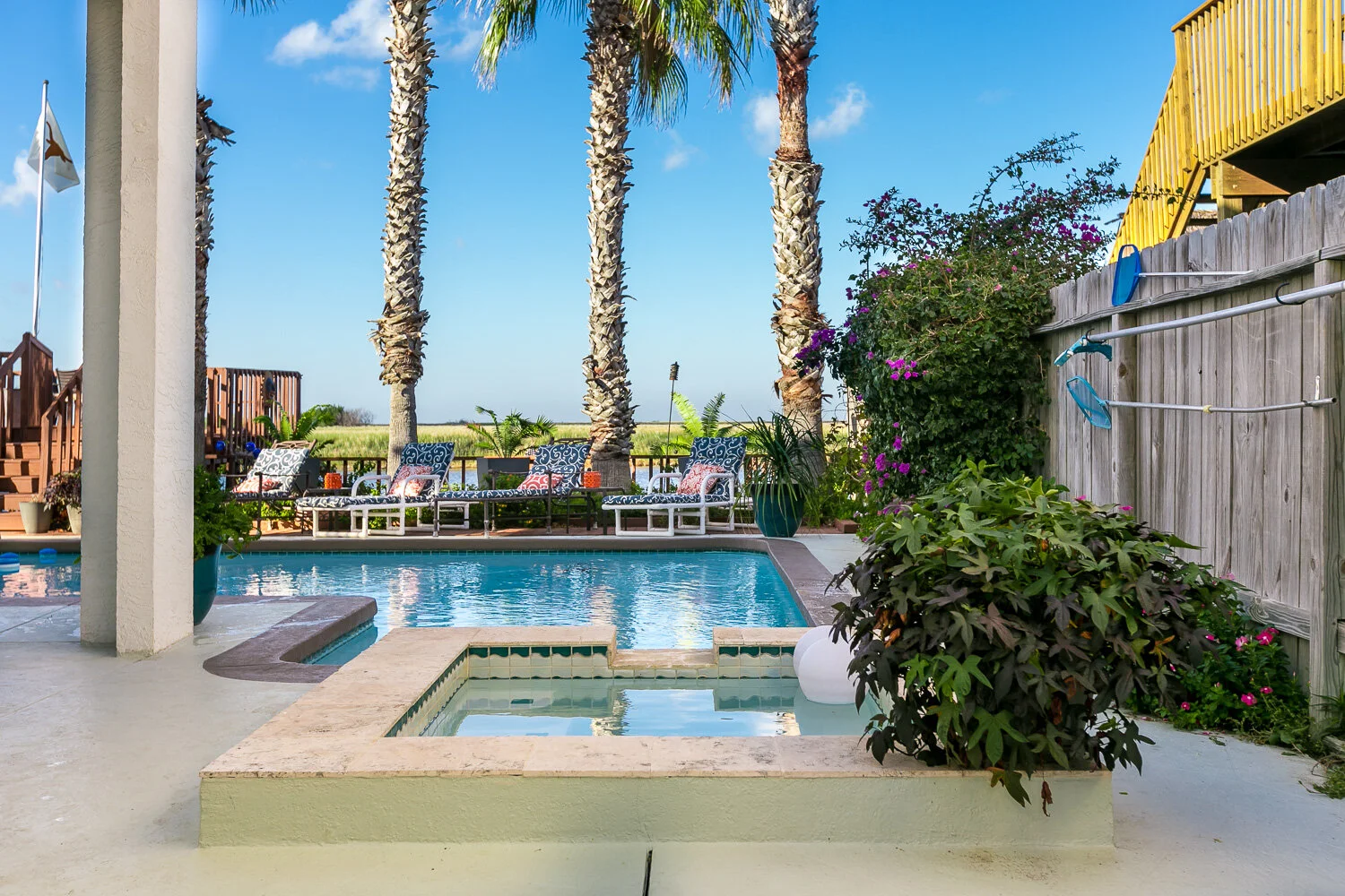 Residential backyard with a swimming pool, palm trees, potted plants, lounge chairs with cushions, and a wooden fence with outdoor shower attachments, under a blue sky.