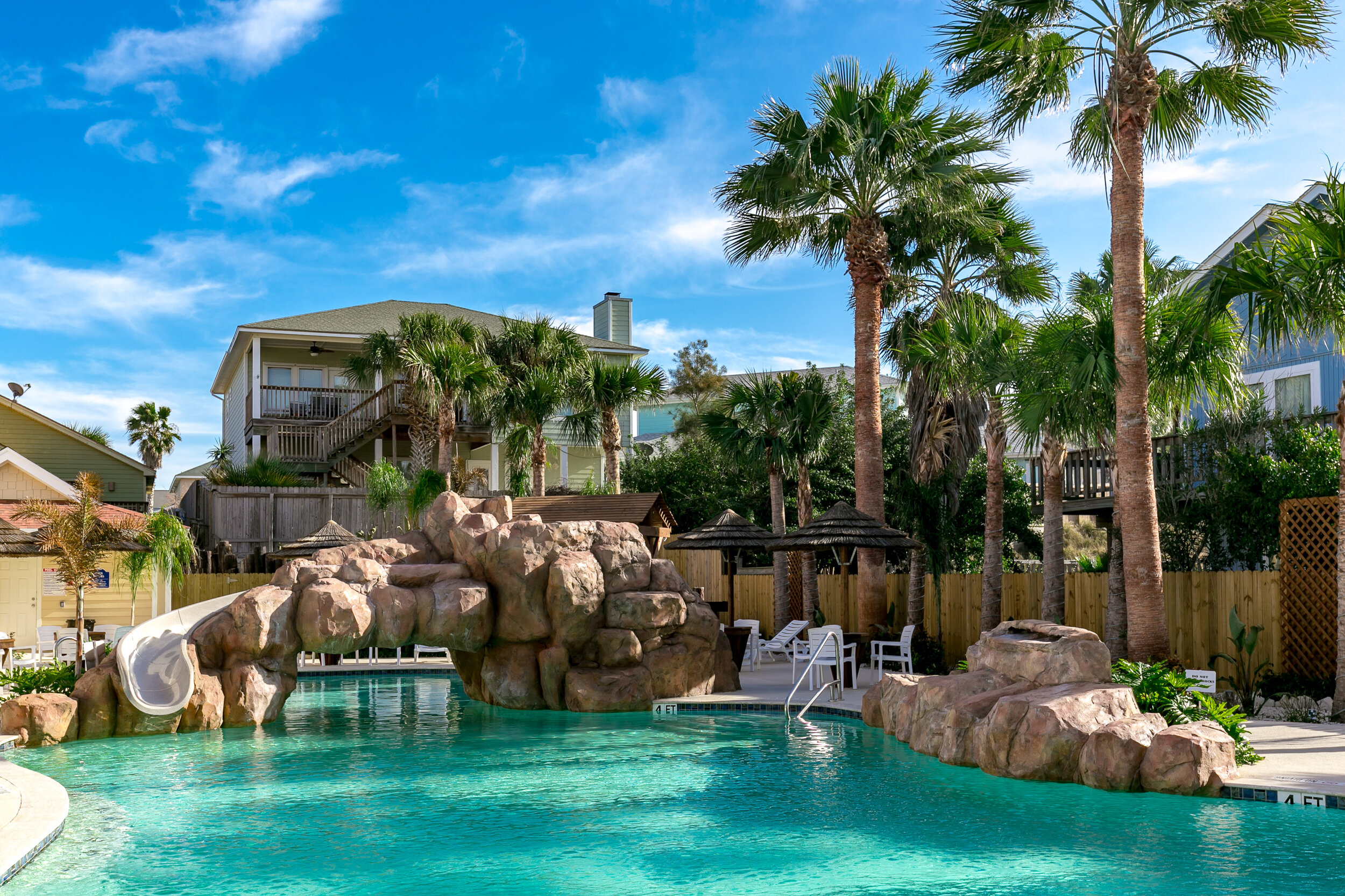 Swimming pool with artificial rock slide, surrounded by palm trees, lounge chairs, and small wooden cabanas, with houses in the background on a sunny day.