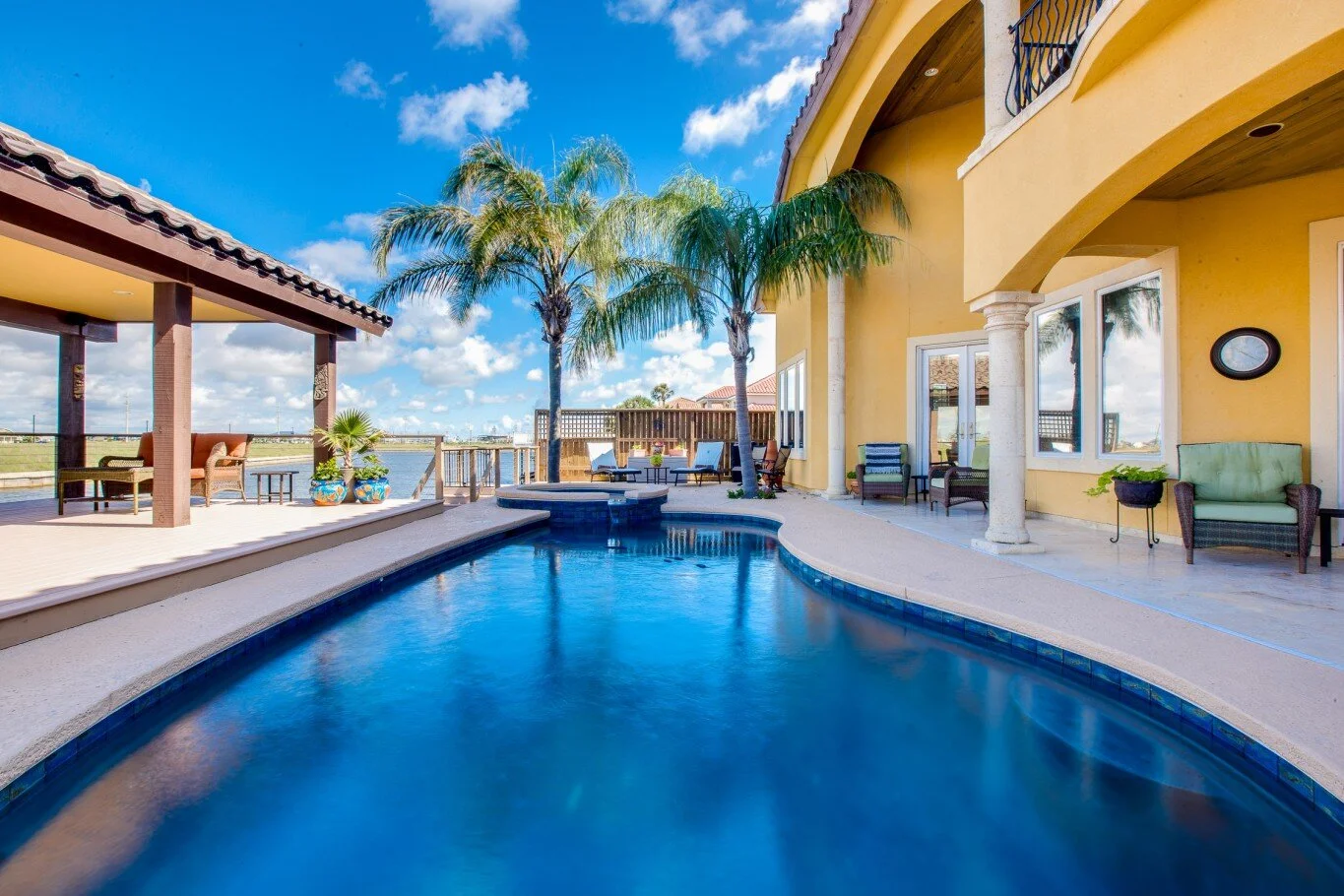 Luxury house with swimming pool, palm trees, and outdoor seating area, under a bright blue sky with clouds.