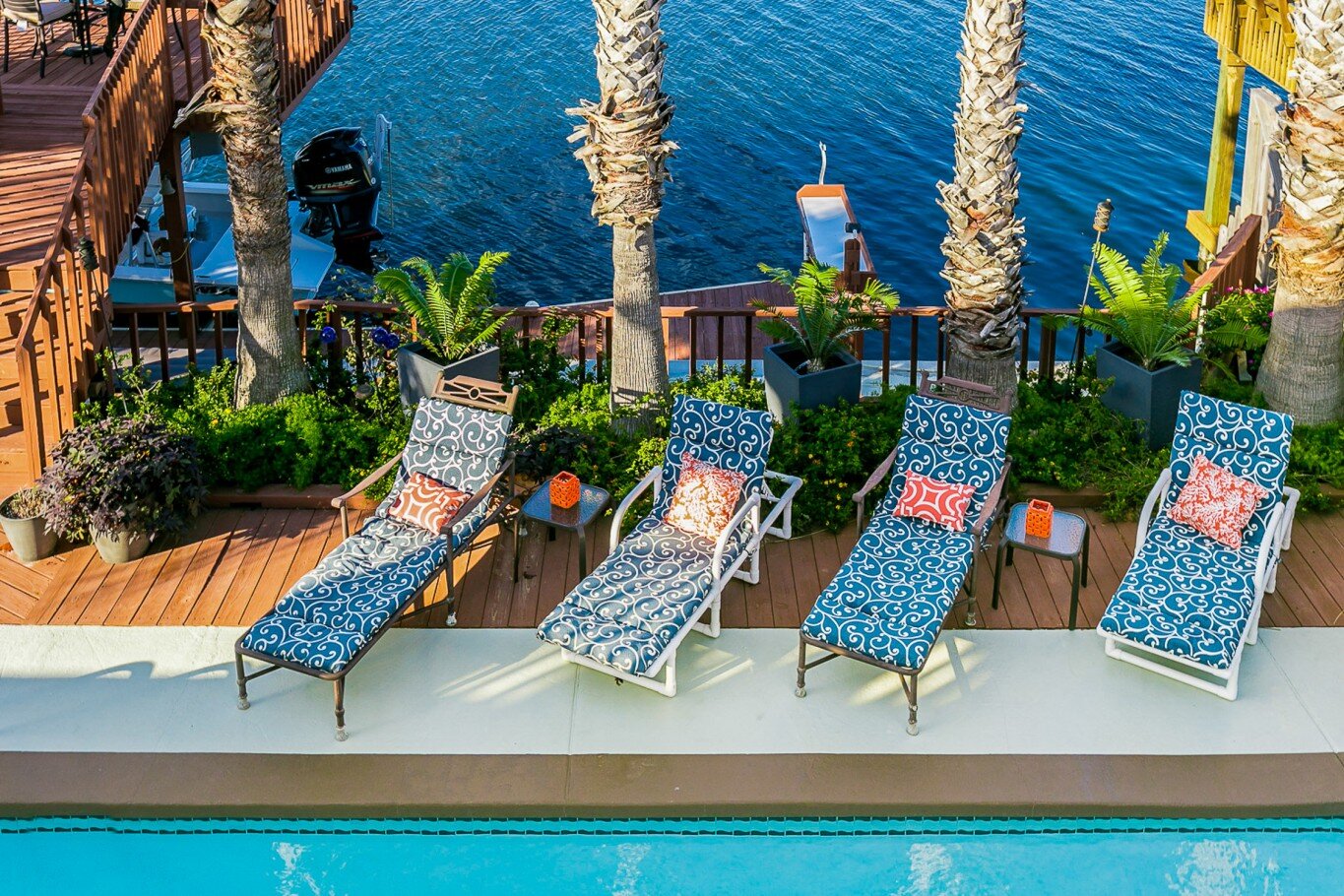 Poolside lounge chairs with patterned cushions, small side tables, and potted plants near a waterfront with palm trees and a boat.