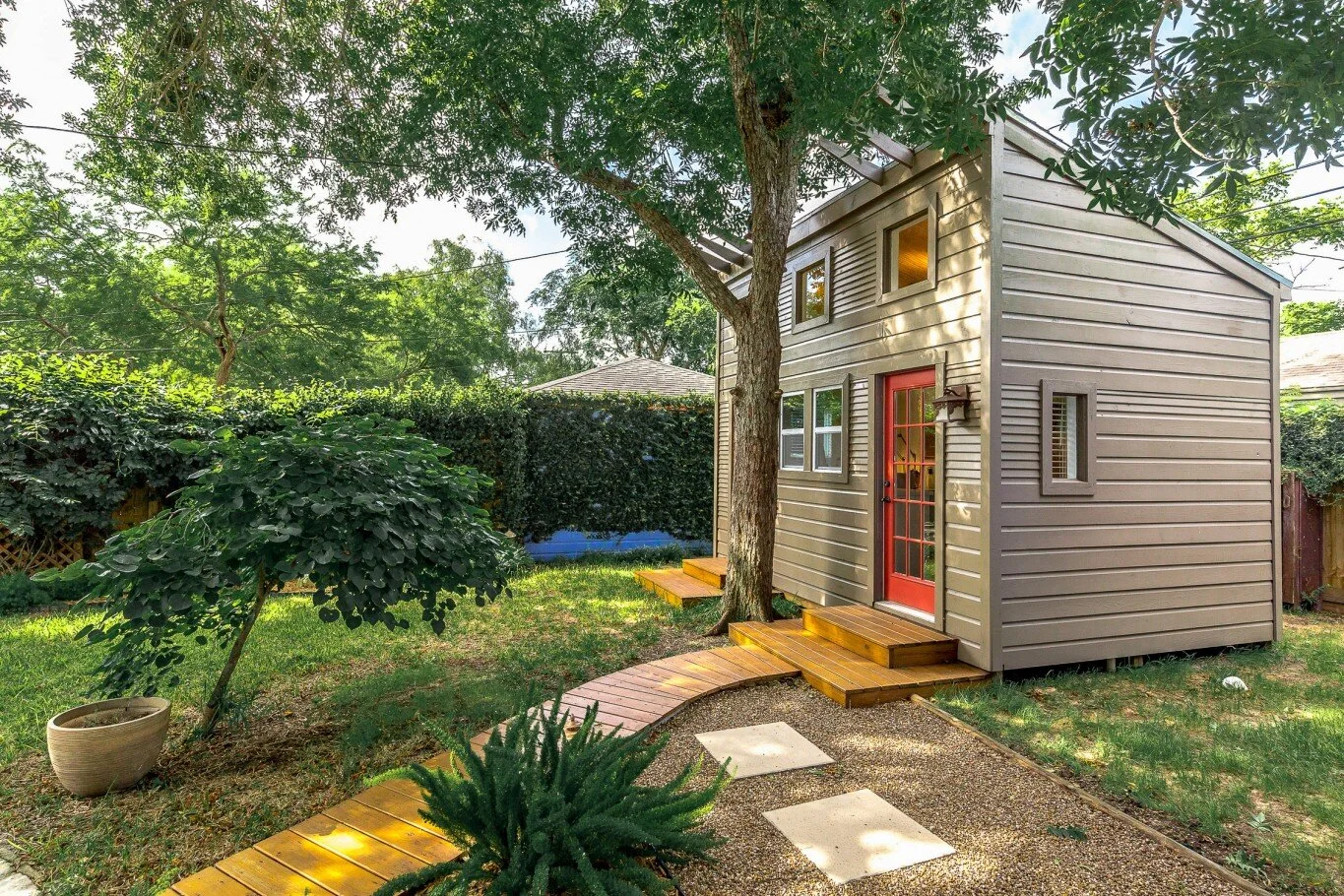 A small, modern two-story house with beige siding, a red front door, and small windows, surrounded by greenery and a garden with a stone pathway.