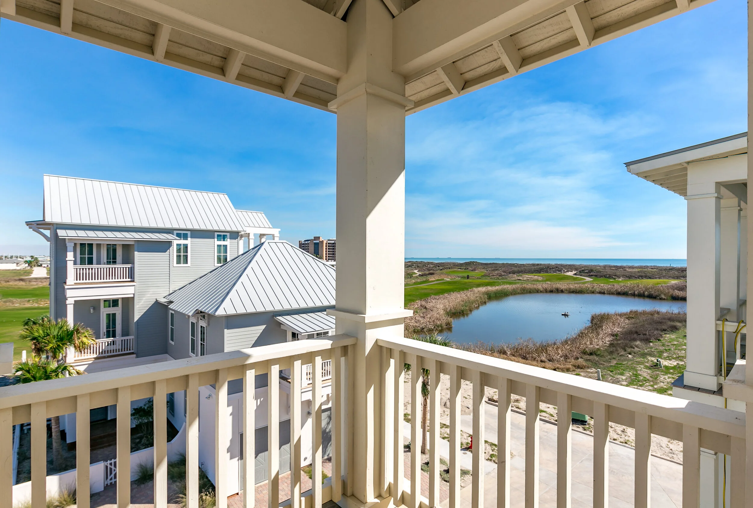 View from a balcony overlooking neighboring beach houses, a pond, and the ocean in the distance under a blue sky.
