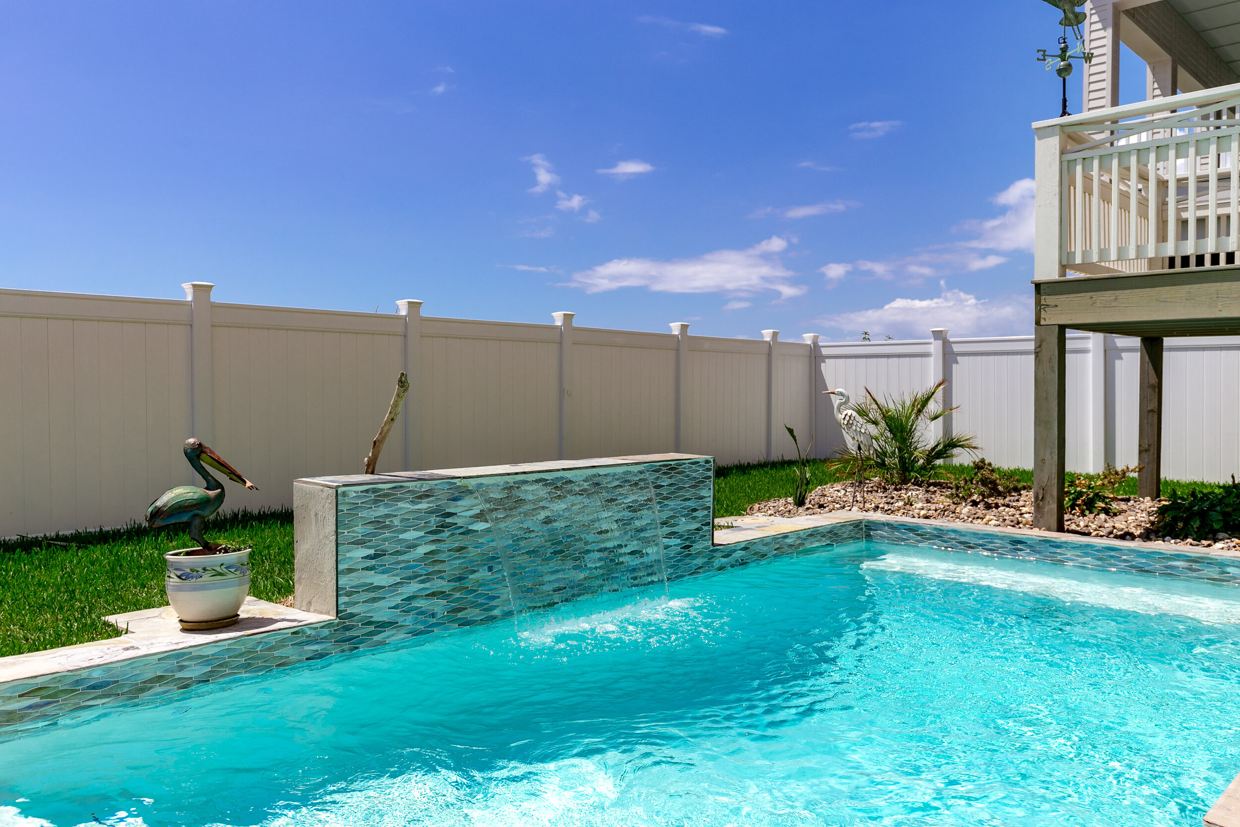 A backyard swimming pool with a blue tiled waterfall feature, textured blue water, white vinyl privacy fence, lawn, decorative bird statues, plants, and a raised deck.