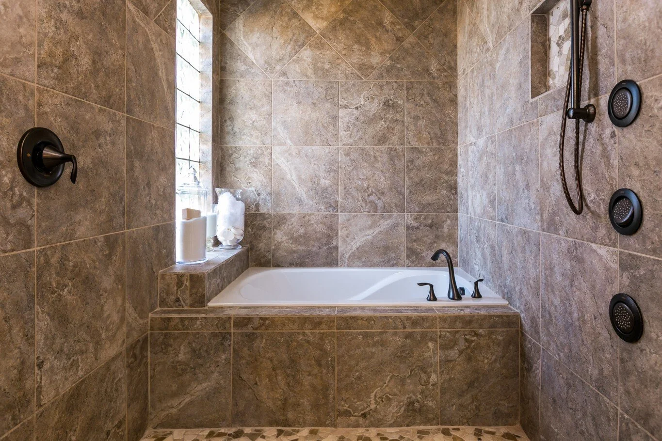 Bathroom shower area with brown tiled walls, a white bathtub, black fixtures, and a small ledge holding candles and toiletries.
