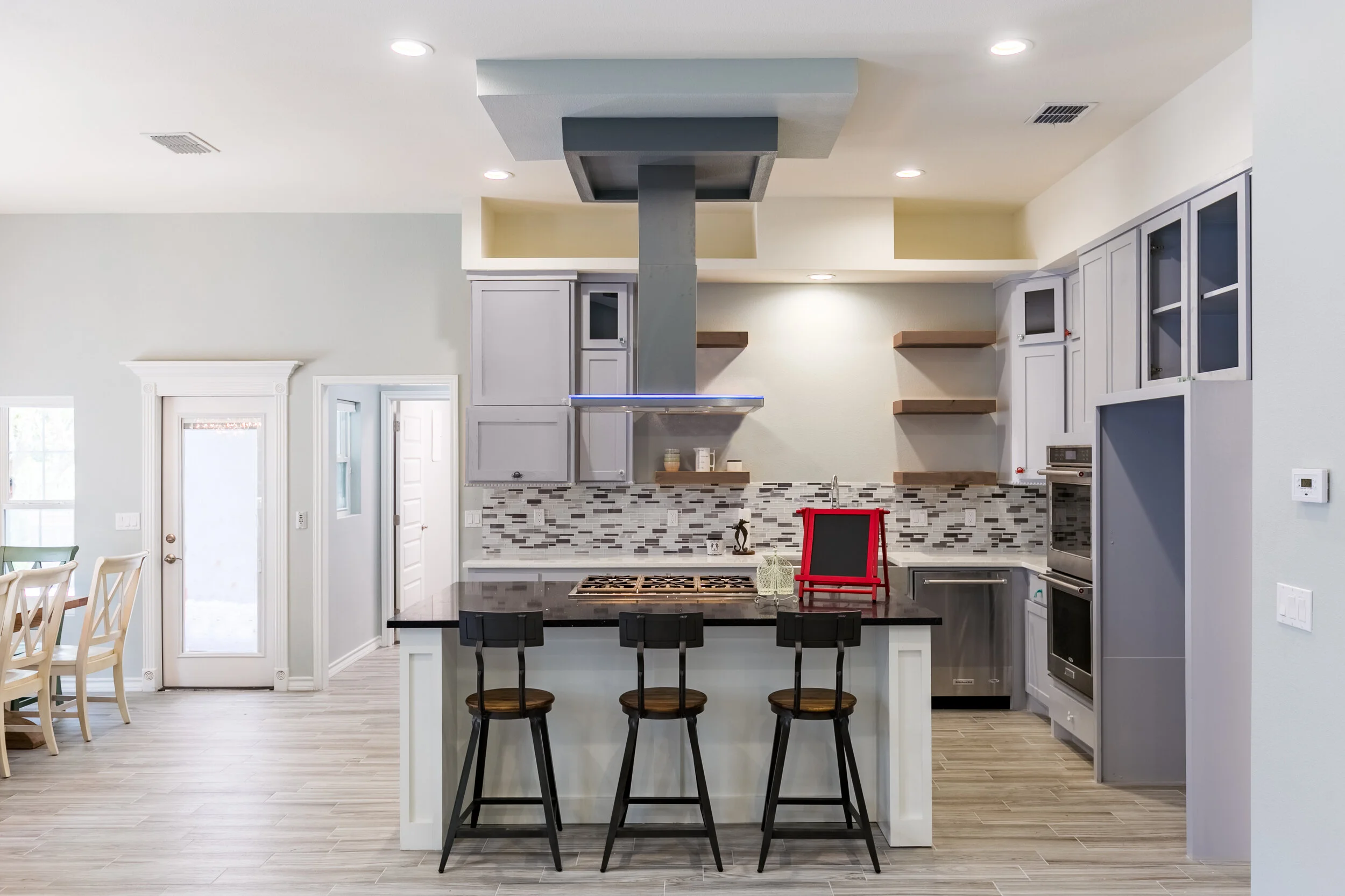 Modern kitchen with gray cabinets, black countertops, and a mosaic tile backsplash. Contains a kitchen island with three black stools, a red chalkboard, and a stainless steel range hood. Open-plan with a dining area visible on the left.
