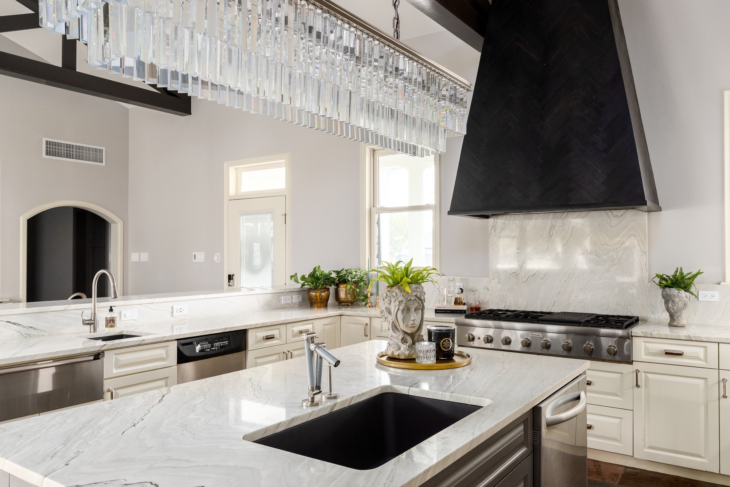 Modern kitchen with white marble countertops and cabinetry, black range hood, plants in decorative pots, large window, crystal chandelier, stainless steel appliances, and a black sink.