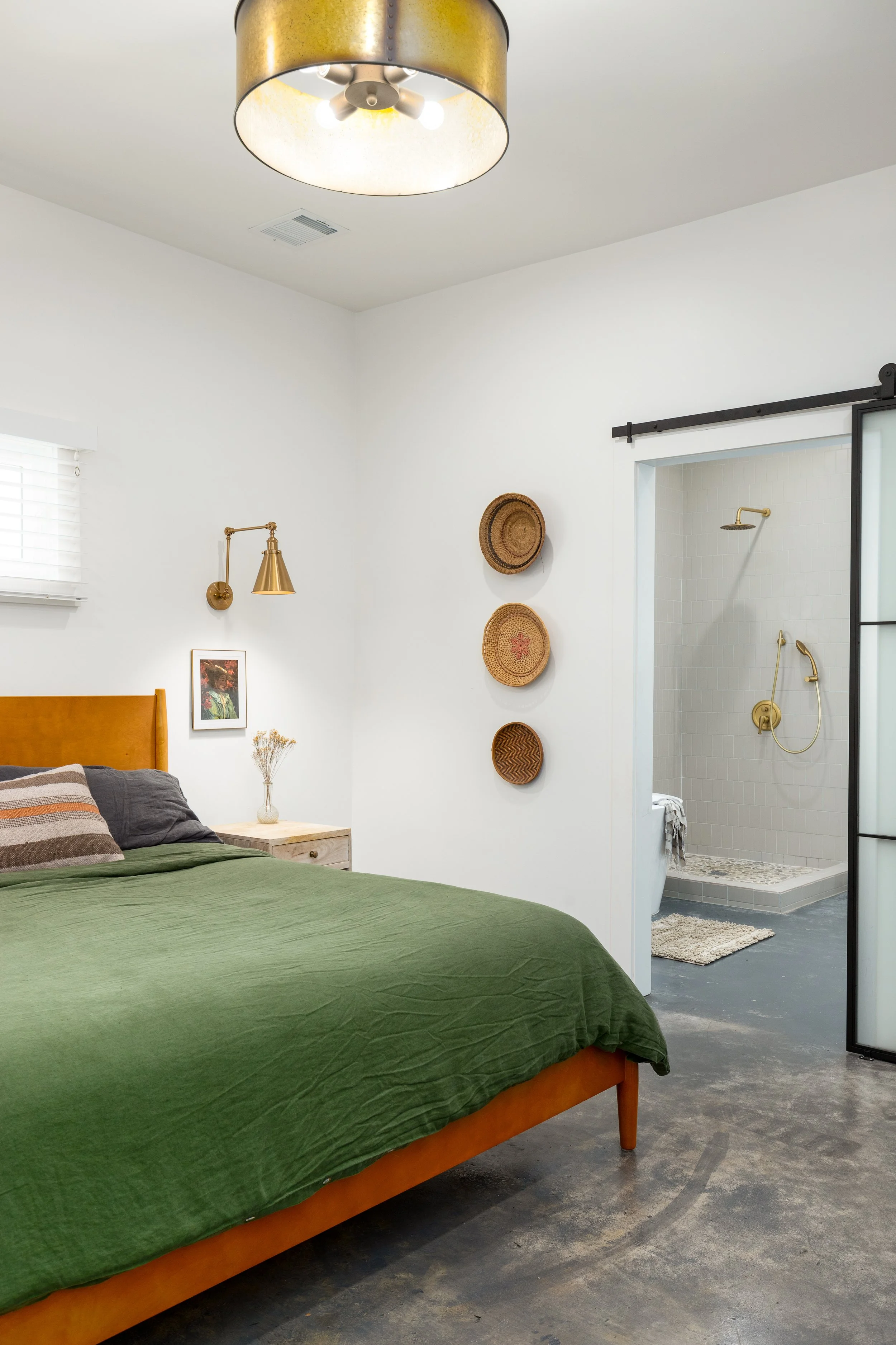 A bedroom with a bed covered in green bedding, wooden headboard, and pillows. A bedside table with a vase of dried flowers. Wall art and wicker baskets on the wall. A ceiling light fixture. An adjacent bathroom with a gold showerhead and a sliding barn door.