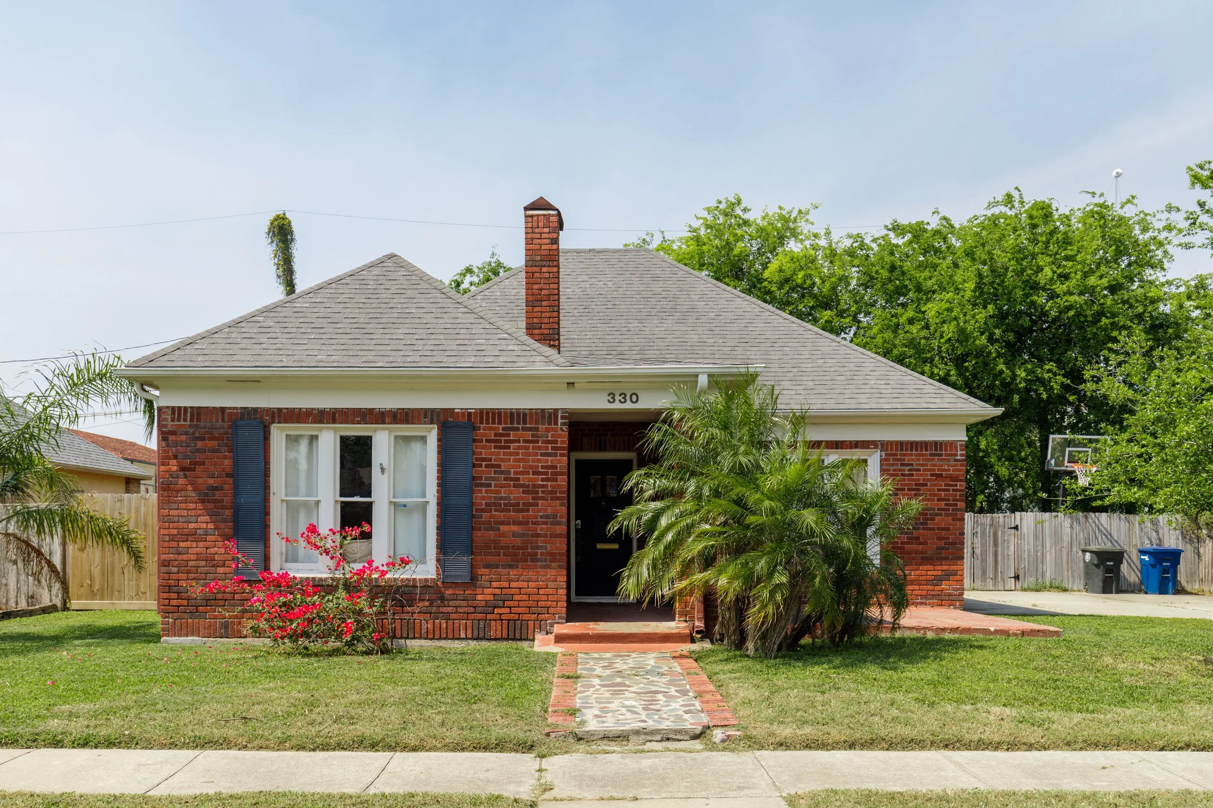 A brick house with a grey shingle roof, a red brick chimney, blue shutters, and a white door. There are pink flowering bushes and green bushes in the front yard, and a sidewalk leading to the front door. A basketball hoop is visible in the background to the right.