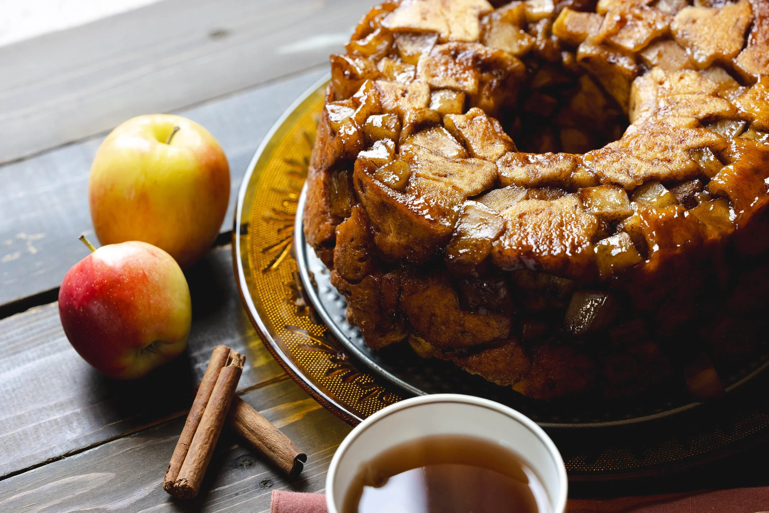 Close-up of a glazed apple cinnamon monkey bread surrounded by apples, cinnamon sticks, and a cup of tea.