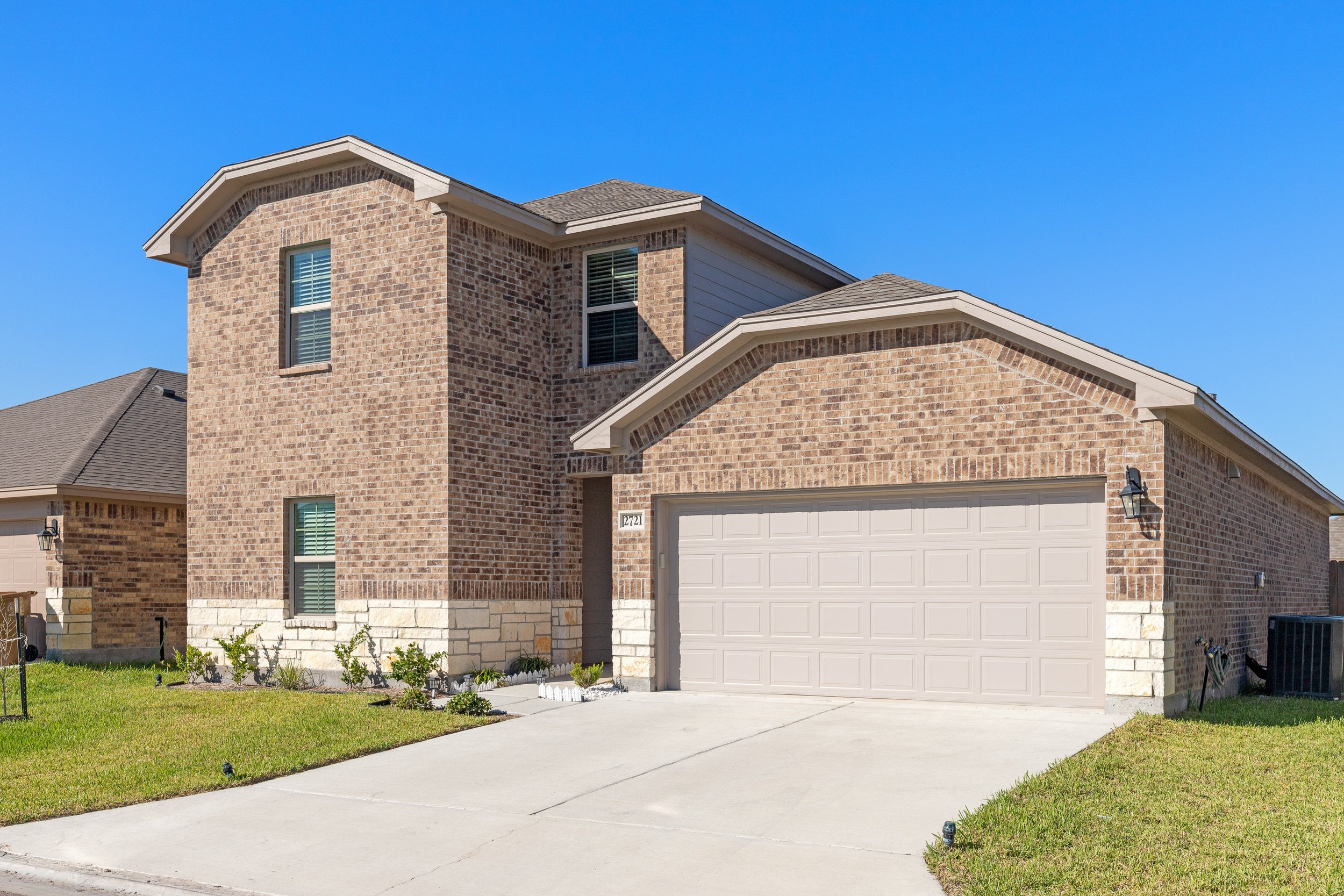 Front view of a two-story house with brick exterior, a two-car garage, a small front garden with grass and shrubs, and a clear blue sky.