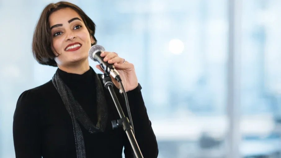 A woman with short brown hair, wearing a black turtleneck and scarf, smiling and holding a microphone in front of a blurred background.