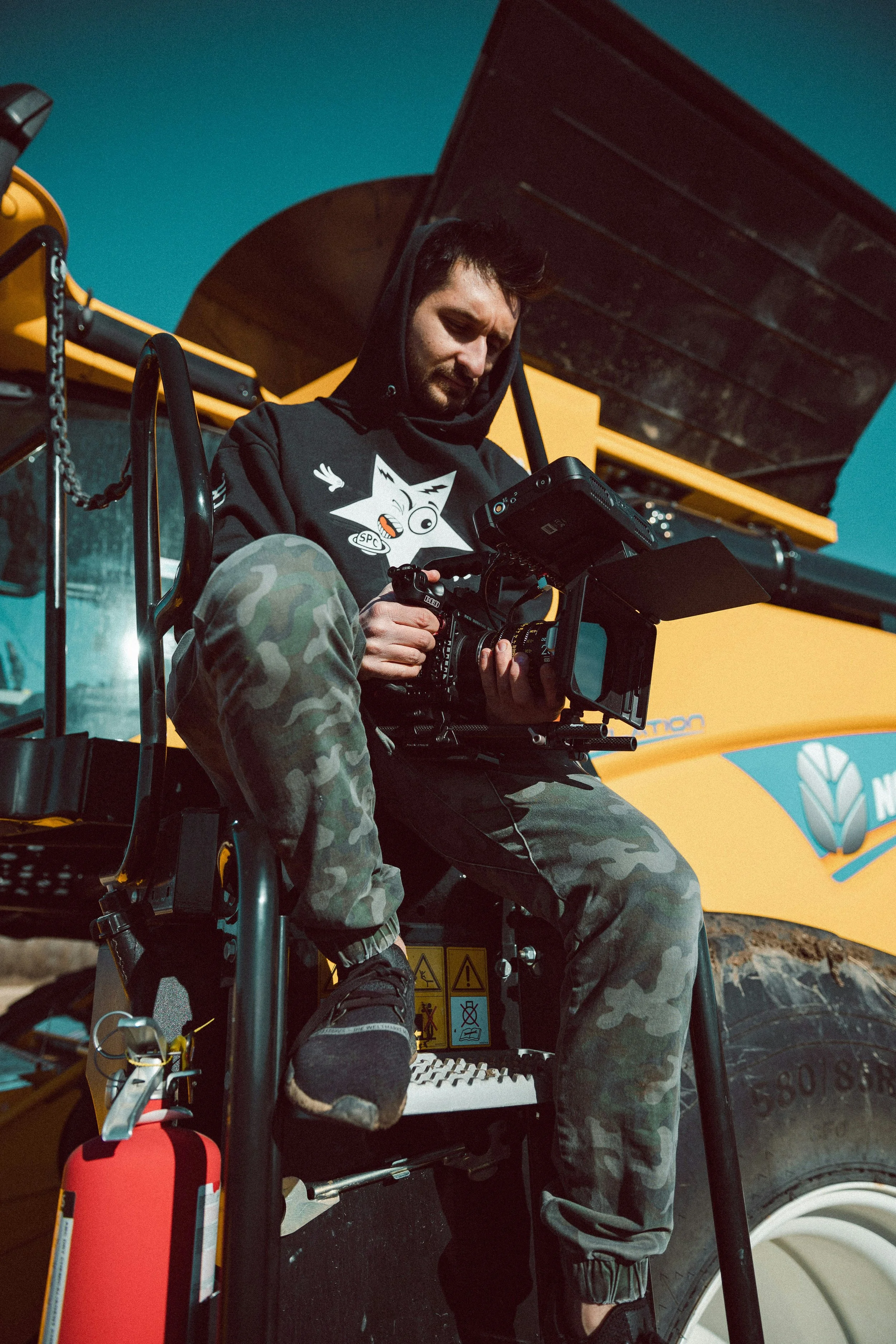 A man in camouflage pants and black shoes sits on a wheelchair, using a professional camera mounted on a tripod for filming. He is outdoors underneath a yellow construction vehicle, with a clear blue sky above.