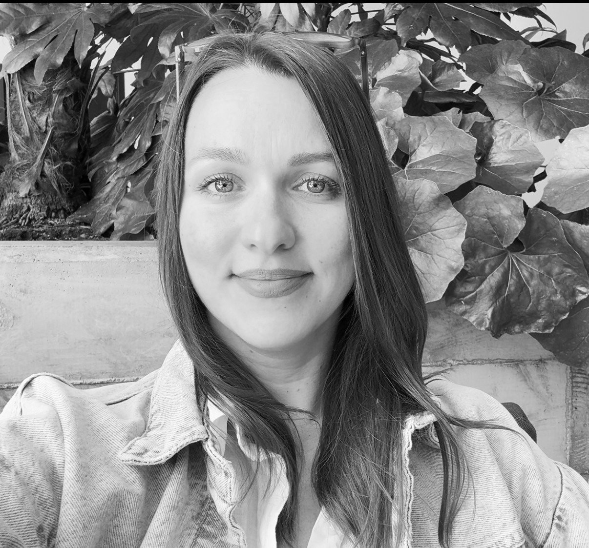 Black and white photo of a woman with straight hair, smiling, sitting in front of leafy plants.