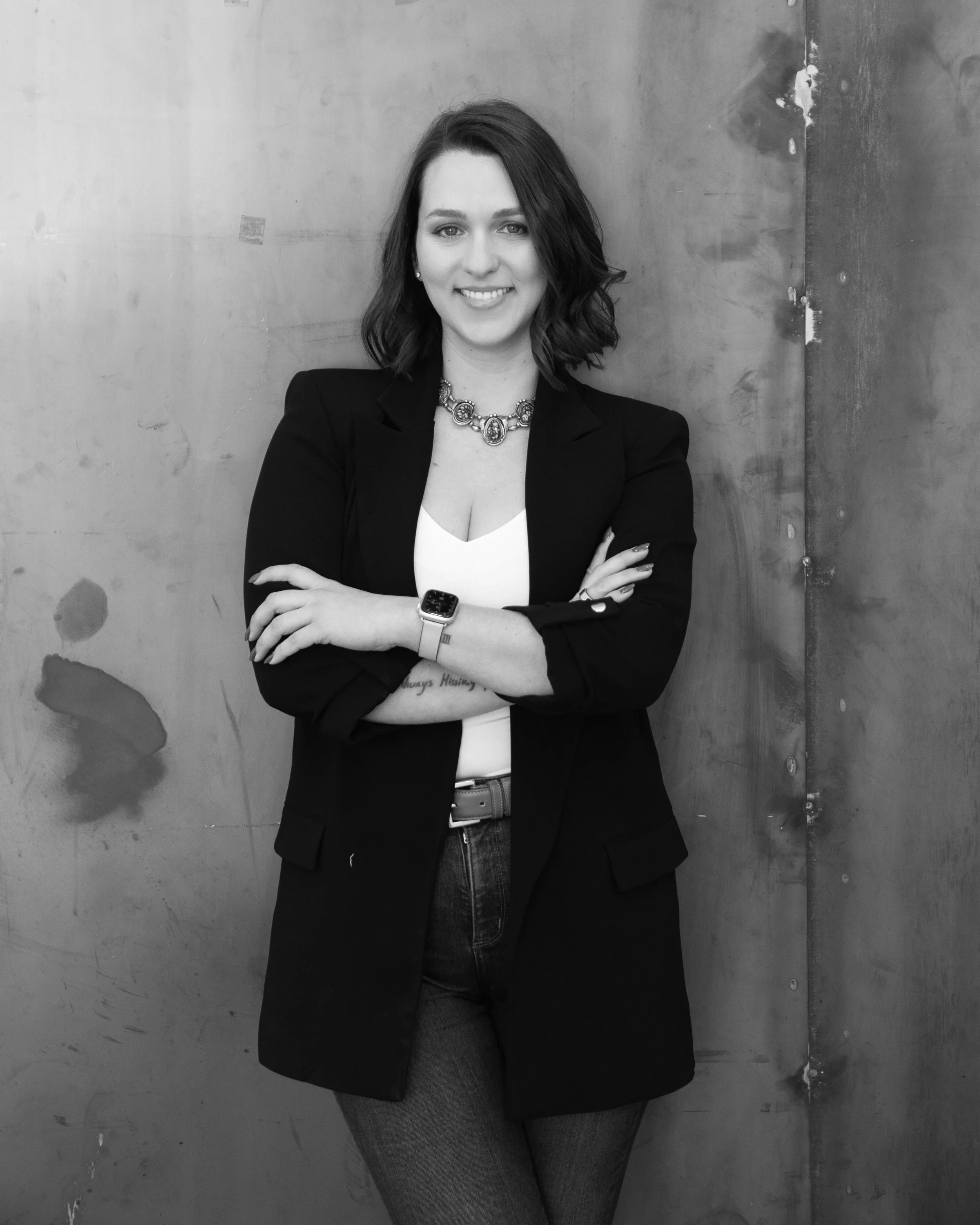 A woman with shoulder-length wavy hair smiling, wearing a black blazer, white top, jeans, a necklace, and a smartwatch, standing with arms crossed against a plain wall.
