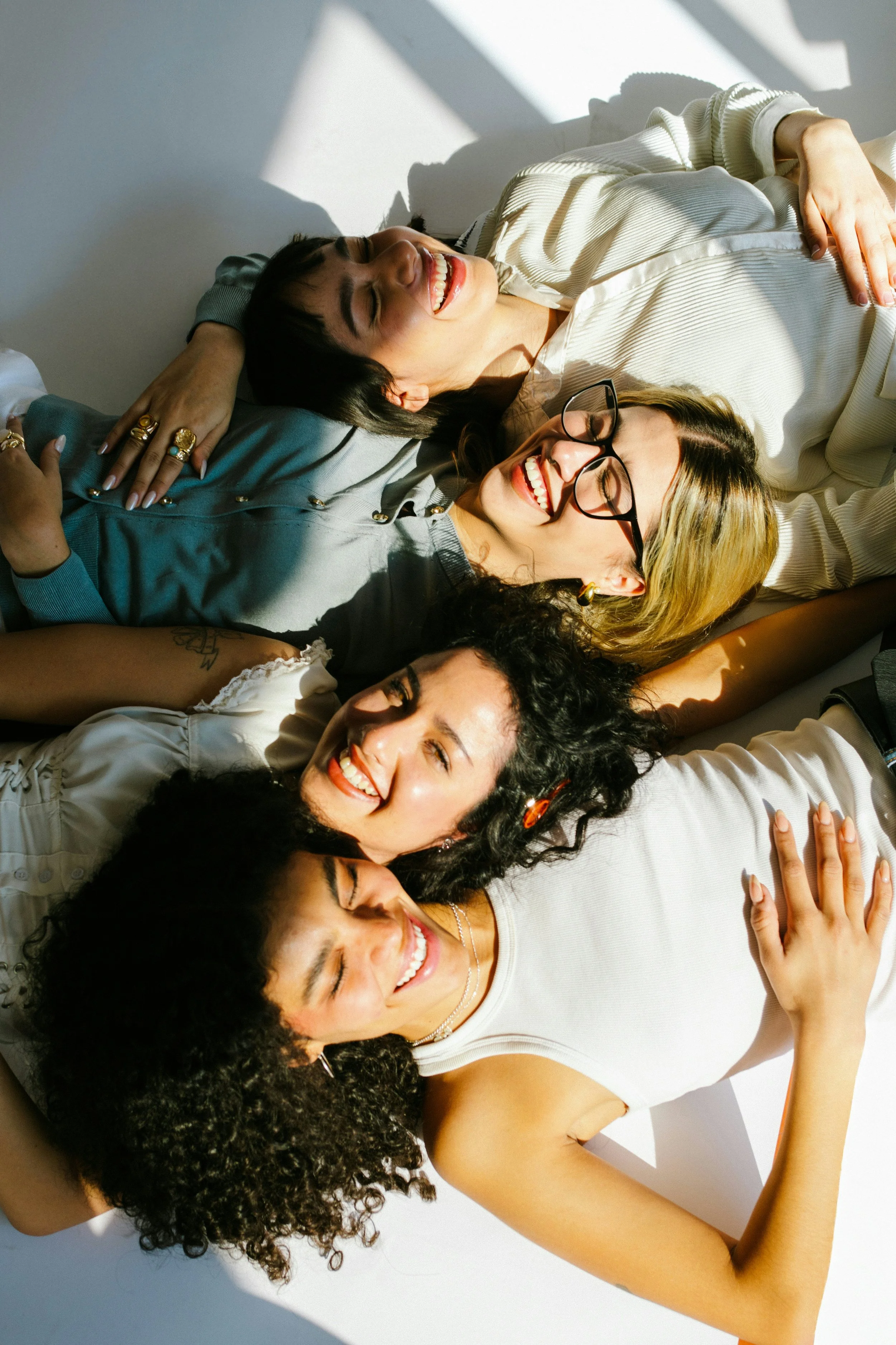 Group of women lying on the floor, smiling and having fun. They are close together, with arms around each other, in a brightly lit space with shadows on the white floor.
