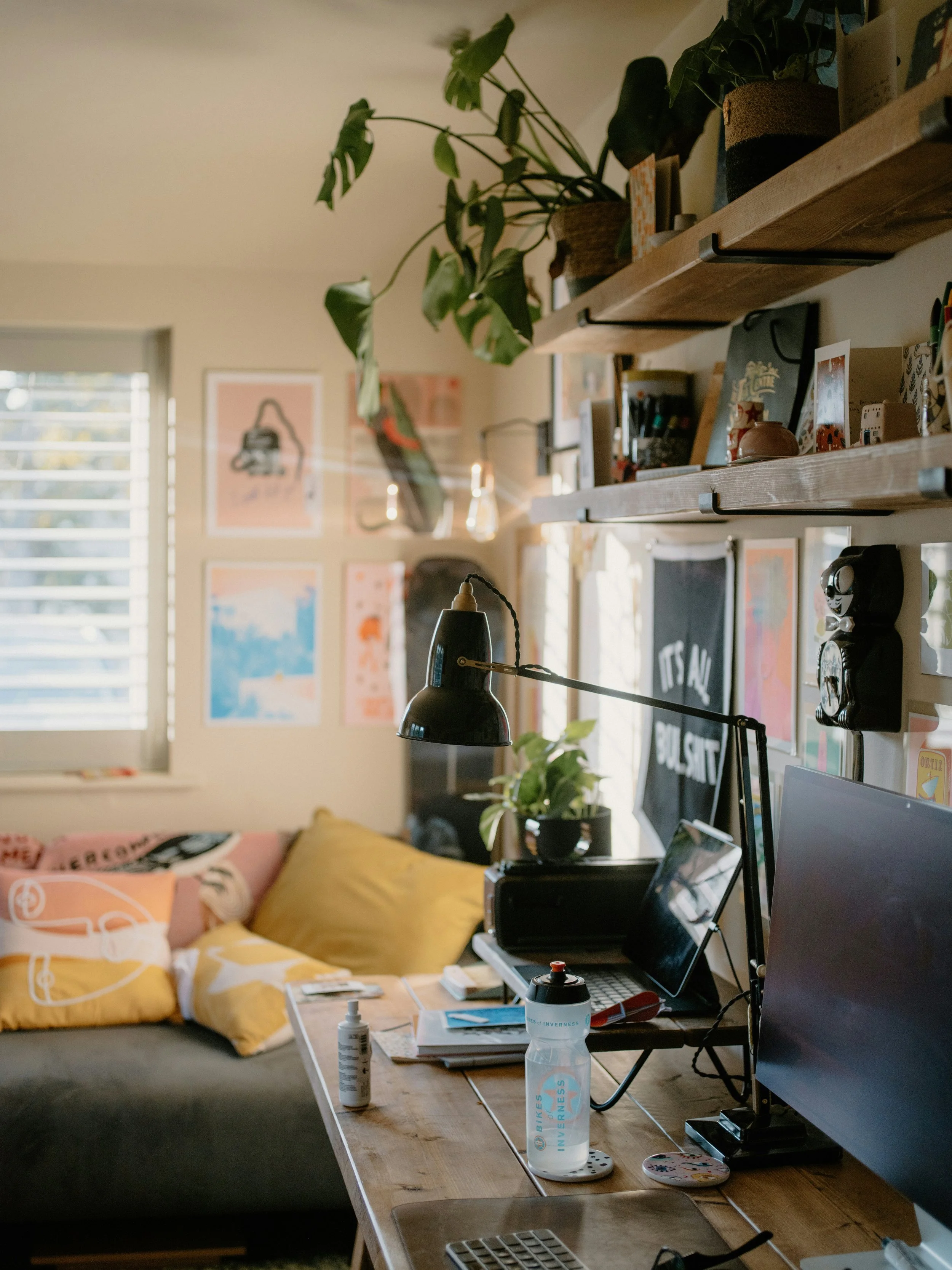 A cozy living room with a wooden desk holding two computer monitors, a laptop, and various office supplies. Behind the desk, there's a comfortable couch with yellow and pink pillows, a wall decorated with colorful art prints, and a large window with blinds providing natural light. The space is decorated with potted plants and personal items, creating a warm and inviting atmosphere.