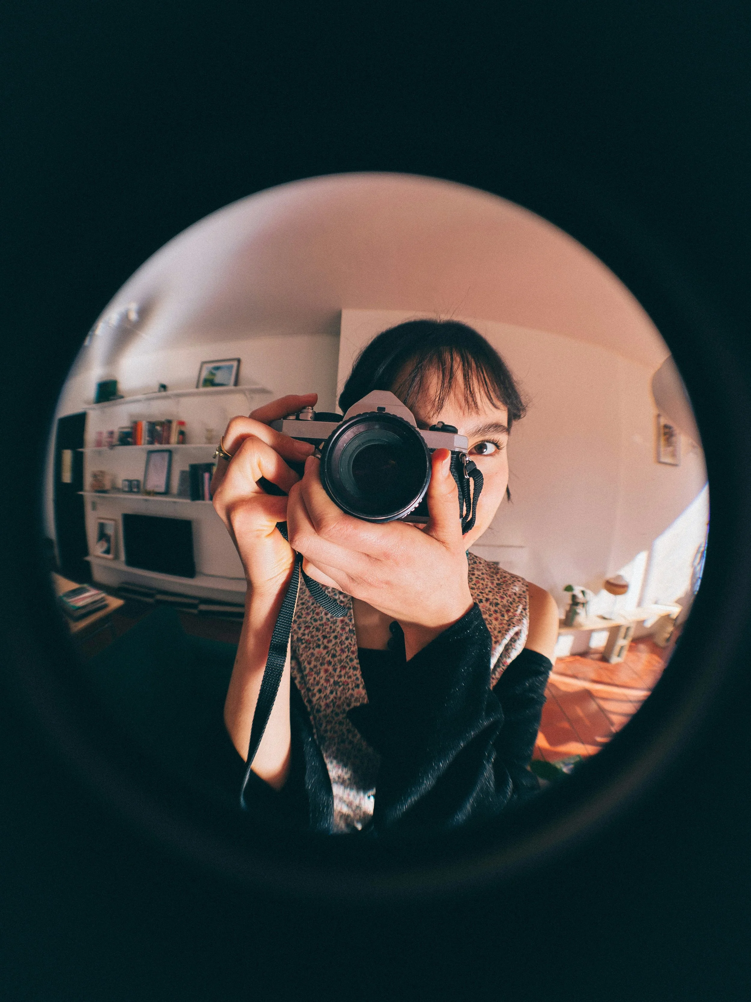 Person taking a selfie in a mirror with a camera, visible eye and part of face, in a room with shelves and framed pictures.