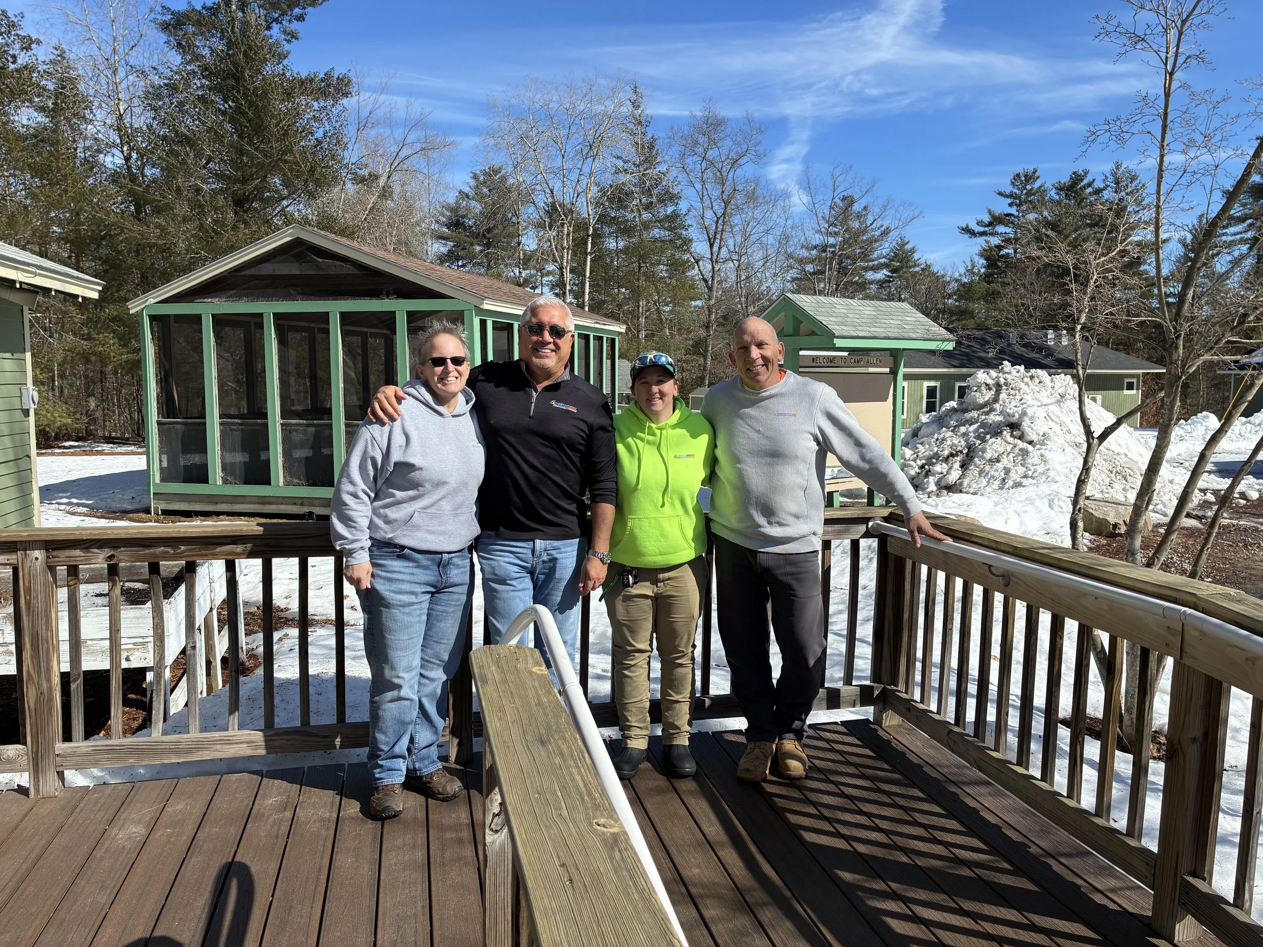 Group of four people standing on deck with long sleeves. Two people with sunglasses; two without. Sunshine with mounds of snow in the background.