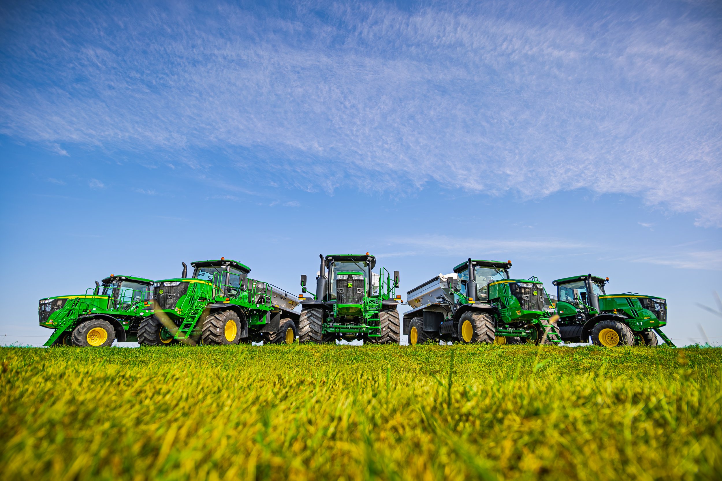 Five green tractors lined up on a grassy field under a partly cloudy blue sky.