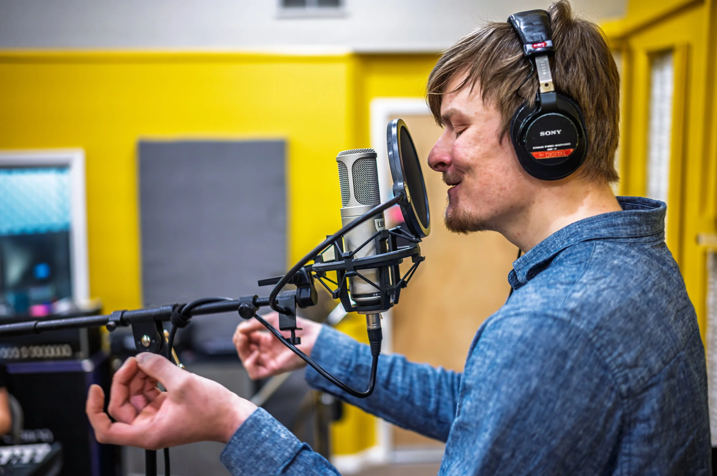 A man with brown hair and a beard wearing headphones in a recording studio, singing into a microphone with a pop filter, while raising his hands in theatrical singing.