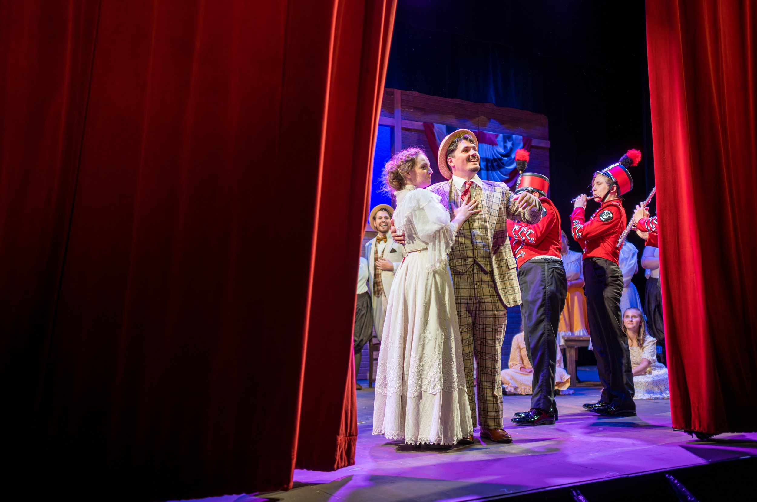 People performing on stage behind red theater curtains, dressed in vintage costumes, with one man smiling and a woman with her hand on his chest, during a theatrical production.
