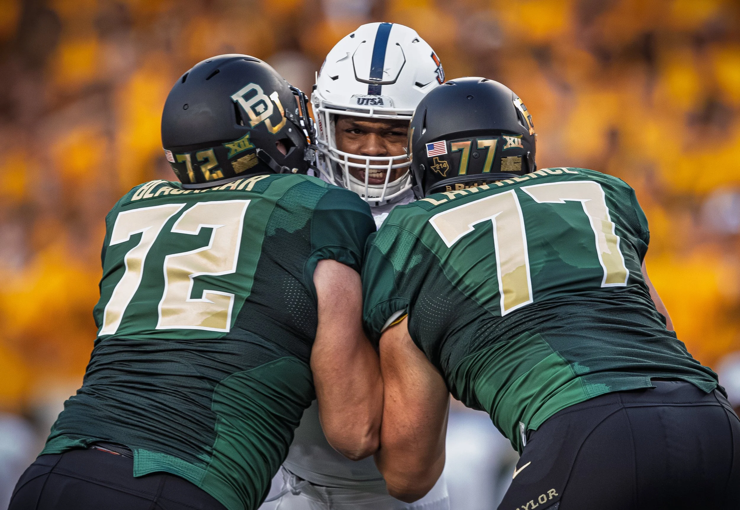 Two football players in green jerseys and black helmets tackling a player in a white jersey with a white helmet during a game.