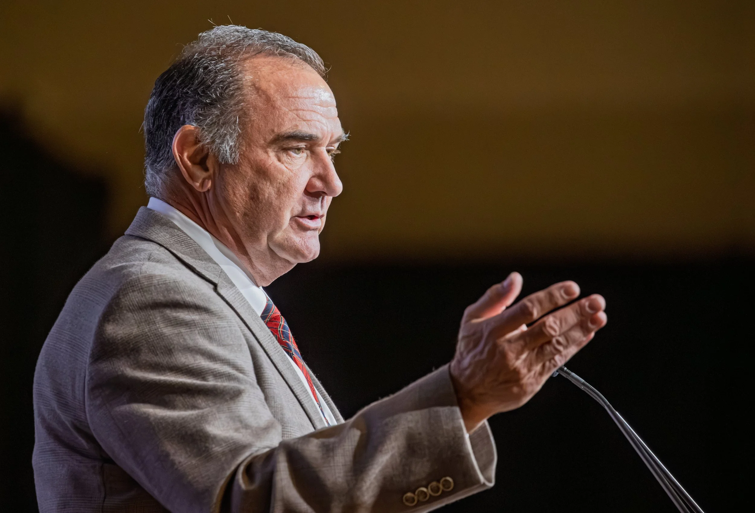 Side profile of a man in a suit speaking at a podium, with a blurred background.