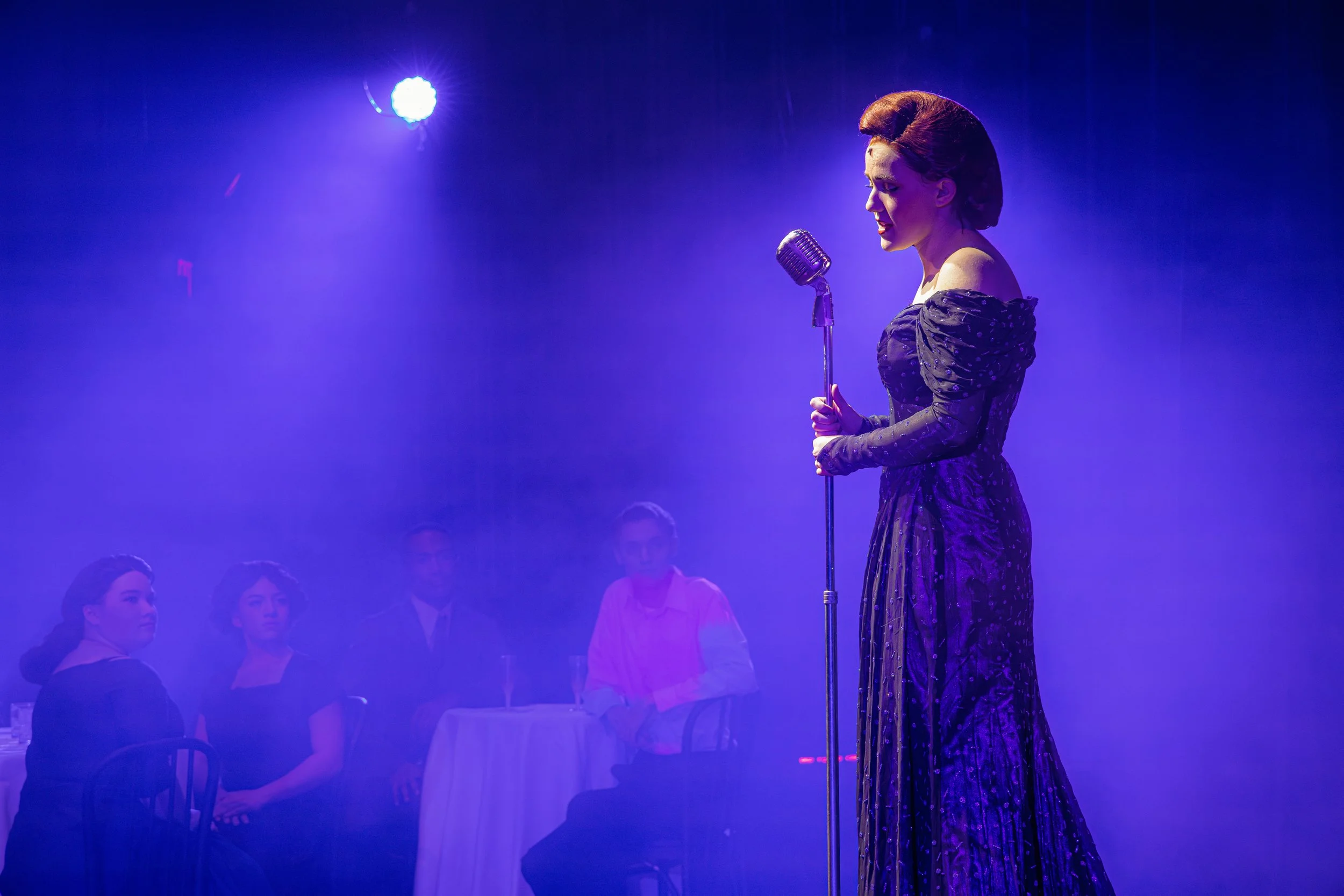 A woman in a vintage dress sings into a microphone on a stage with purple lighting, audience members seated around her.