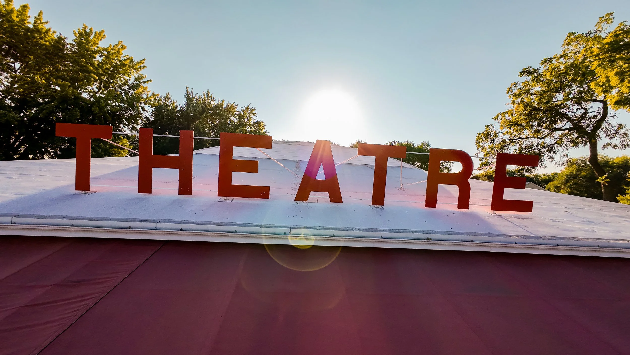 The word 'THEATRE' spelled out with large orange letters attached to a roof, with trees and a clear sky in the background, and sunlight creating a lens flare.
