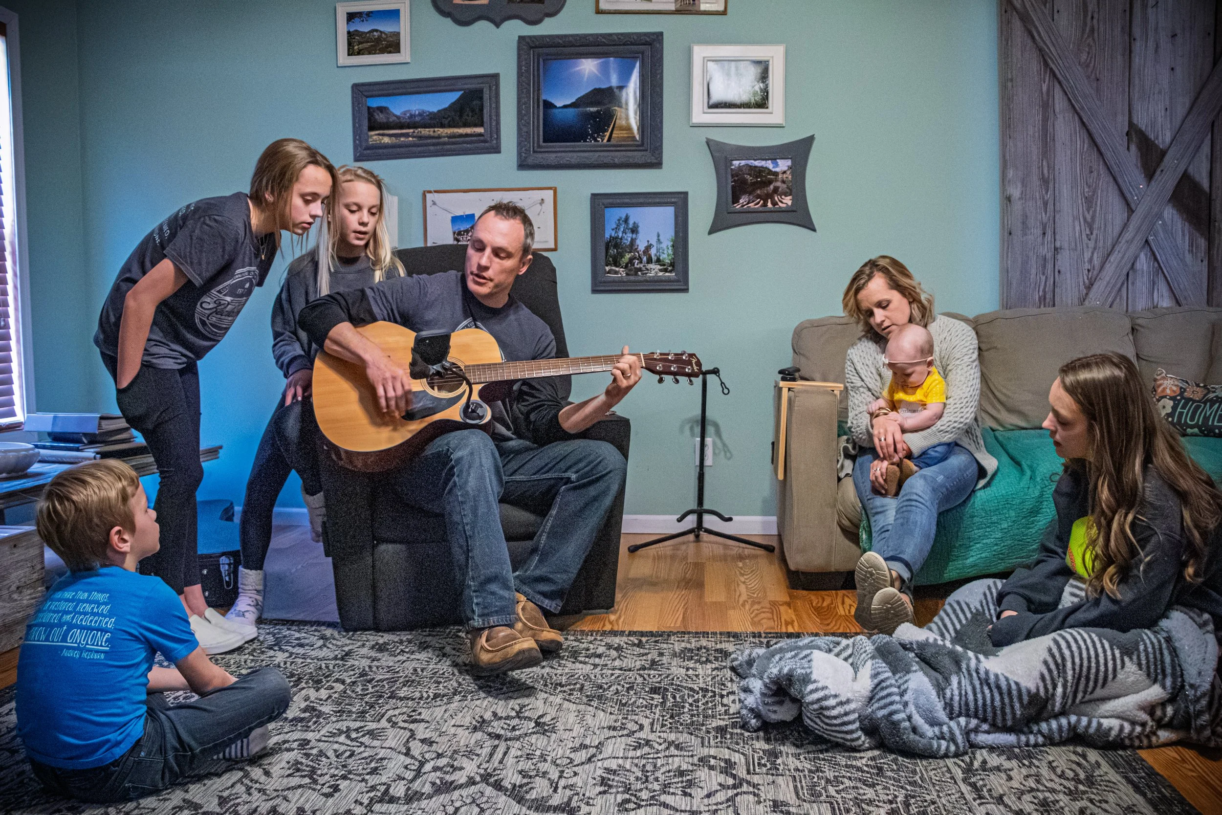 A group of children and adults sitting on the floor and on a sofa in a living room, listening to a man playing an acoustic guitar. The walls are decorated with framed landscape photographs, and the room has a cozy, casual atmosphere.