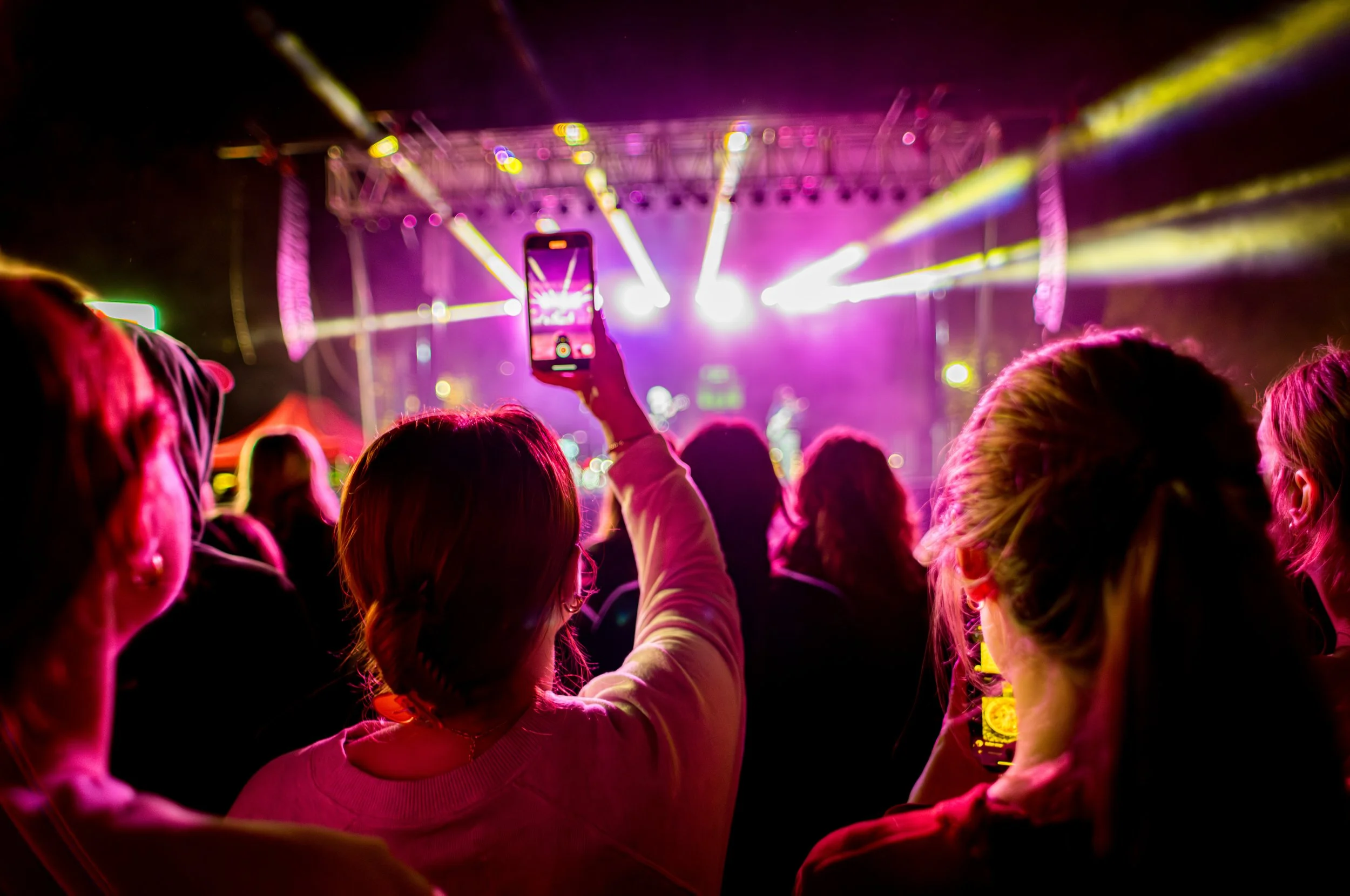 Audience at a concert or festival enjoying colorful stage lights with one person holding up a smartphone to capture the performance.