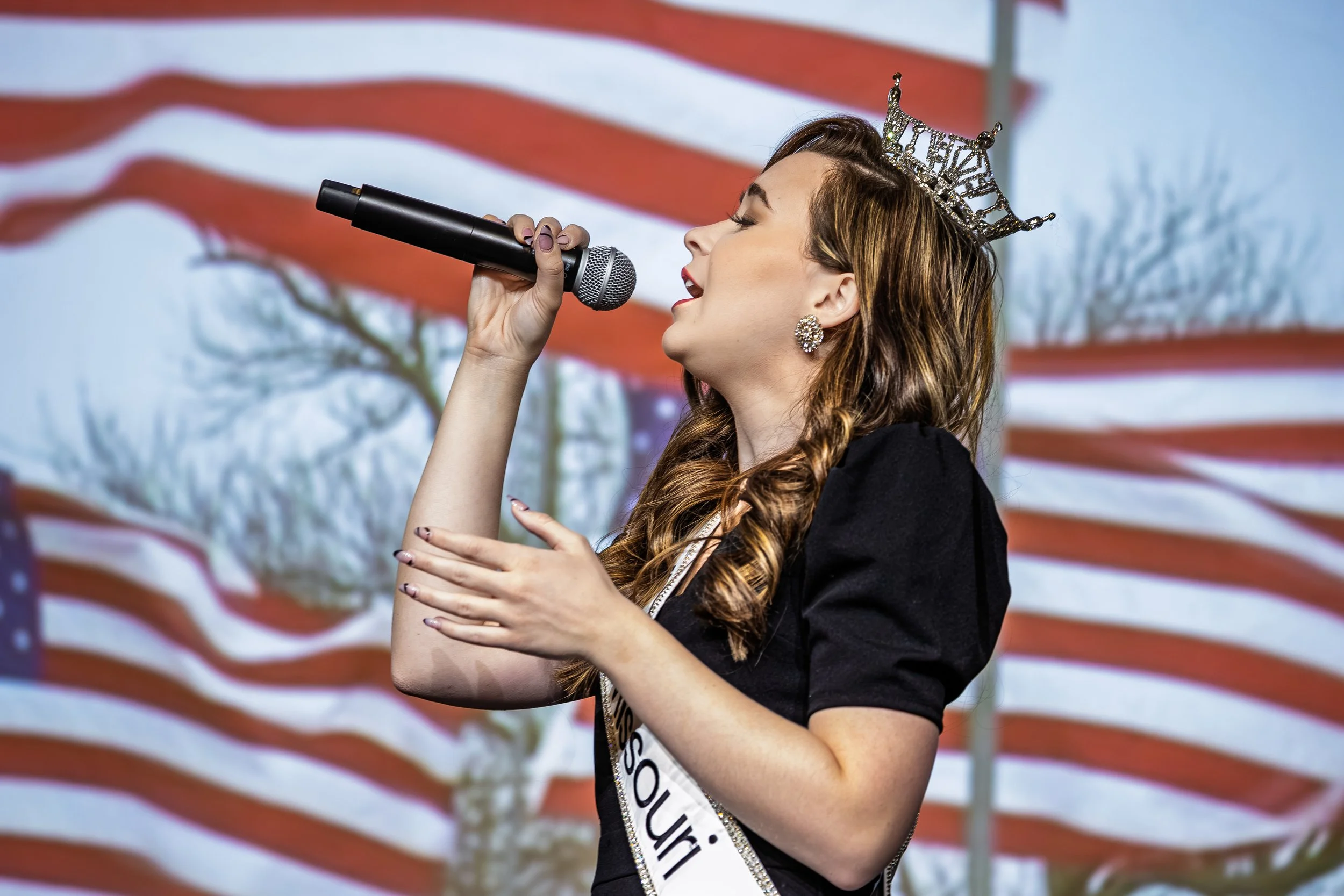 Young woman wearing a crown and earrings, singing into a microphone during patriotic event with American flag in the background.