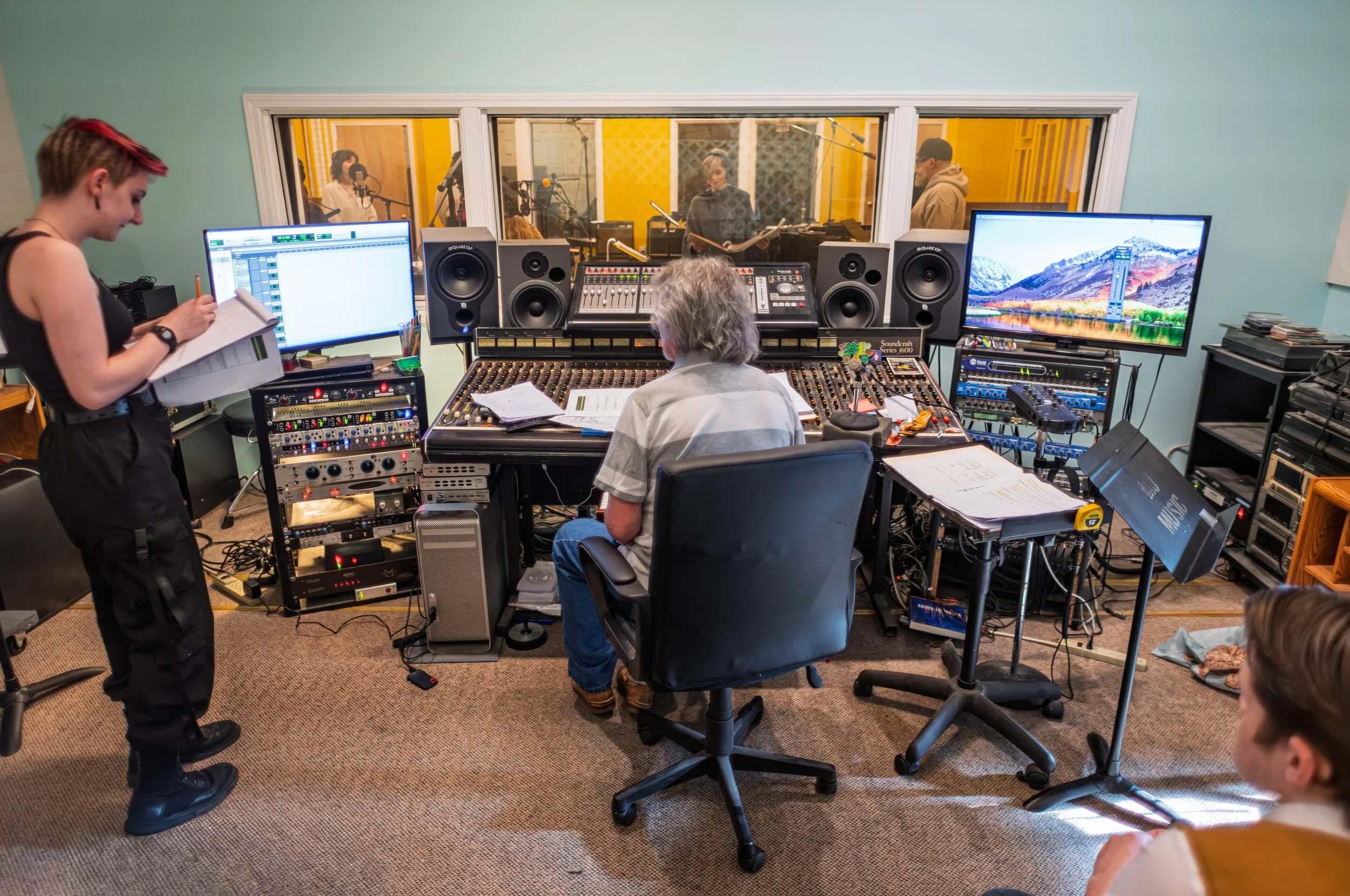 Recording studio control room with two large computer monitors, six speakers, a sound mixer, and staff working while a woman in the background reads in a vocal booth.