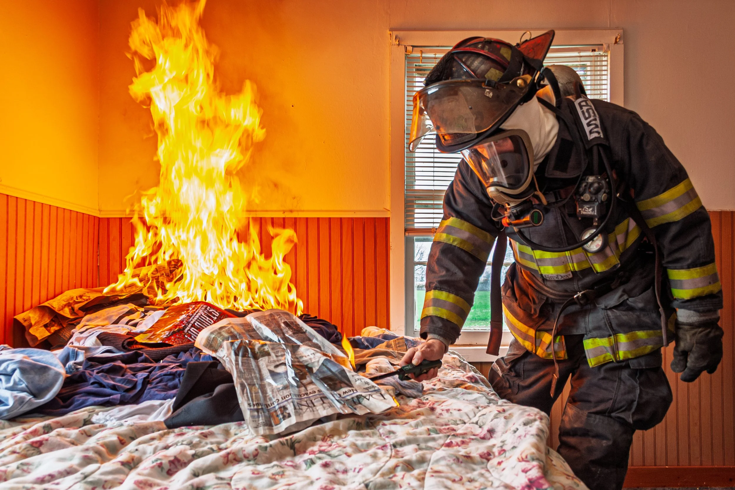 Fire burning on a bed near a firefighter in protective gear.