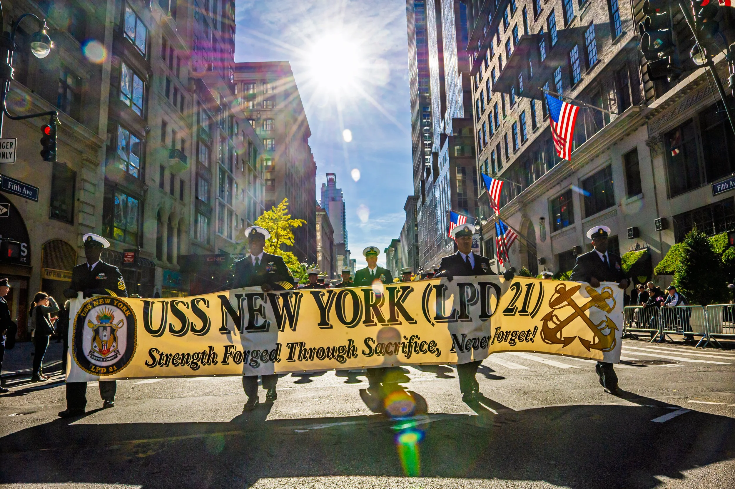 A parade on a sunny day with military personnel marching and carrying a large yellow banner that reads, 'USS NEW YORK (LPD 21)' with symbols of strength and sacrifice. The background features tall city buildings, American flags, and a bright sun with lens flares.