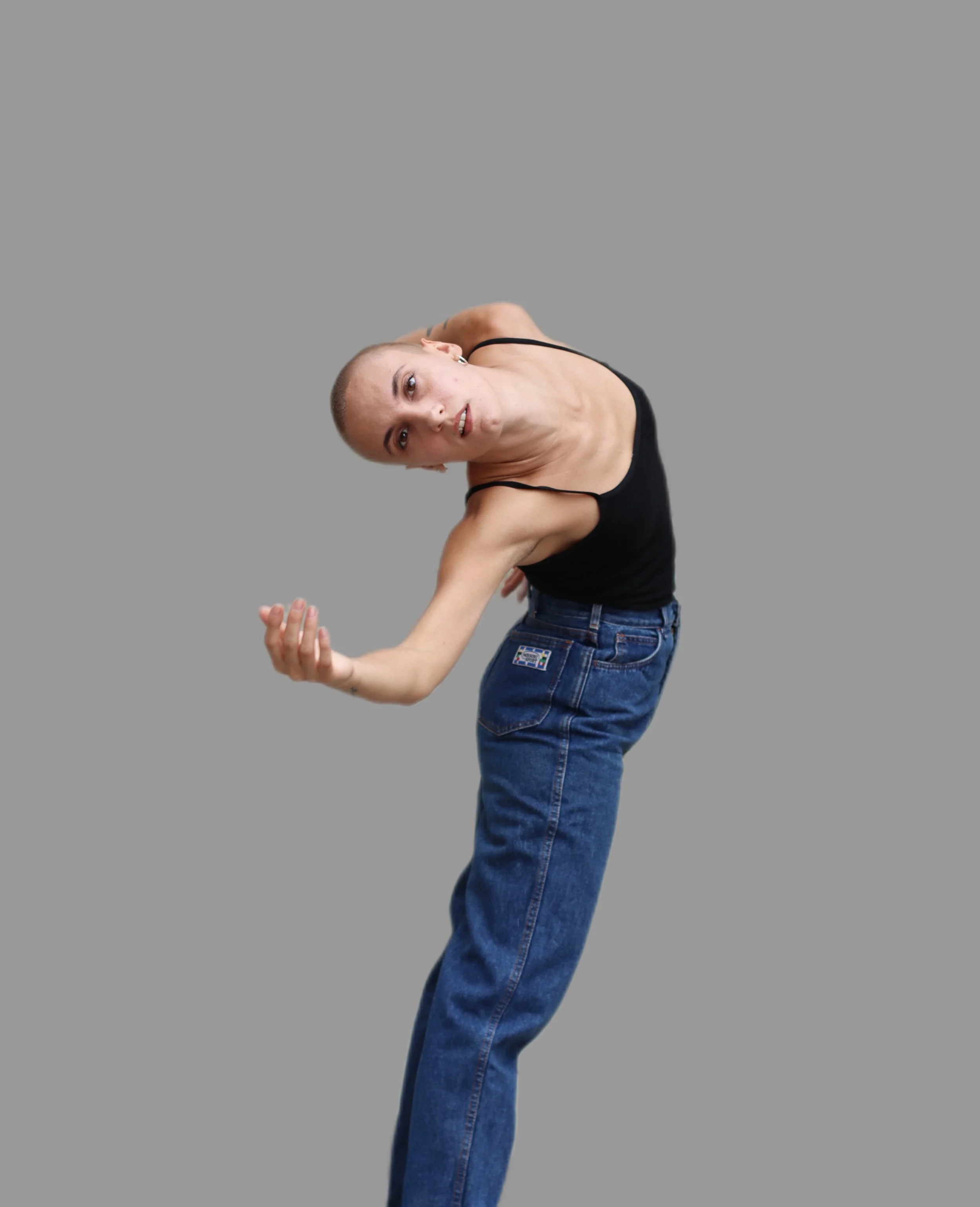 Person with a shaved head wearing a black tank top and blue jeans, leaning to the side against a gray background.