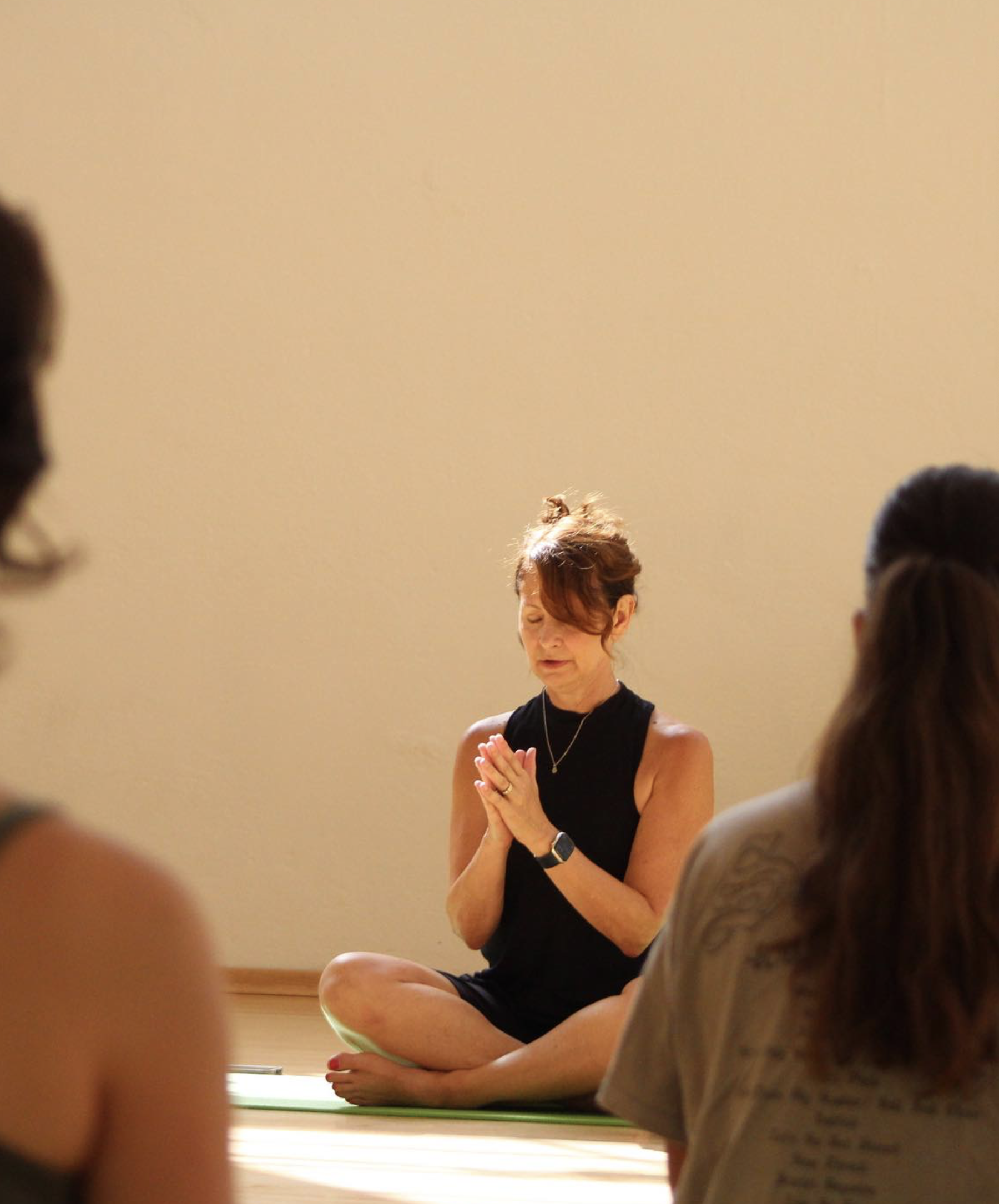 A woman practicing yoga or meditation outdoors, sitting cross-legged with hands in prayer position, surrounded by a group of people.