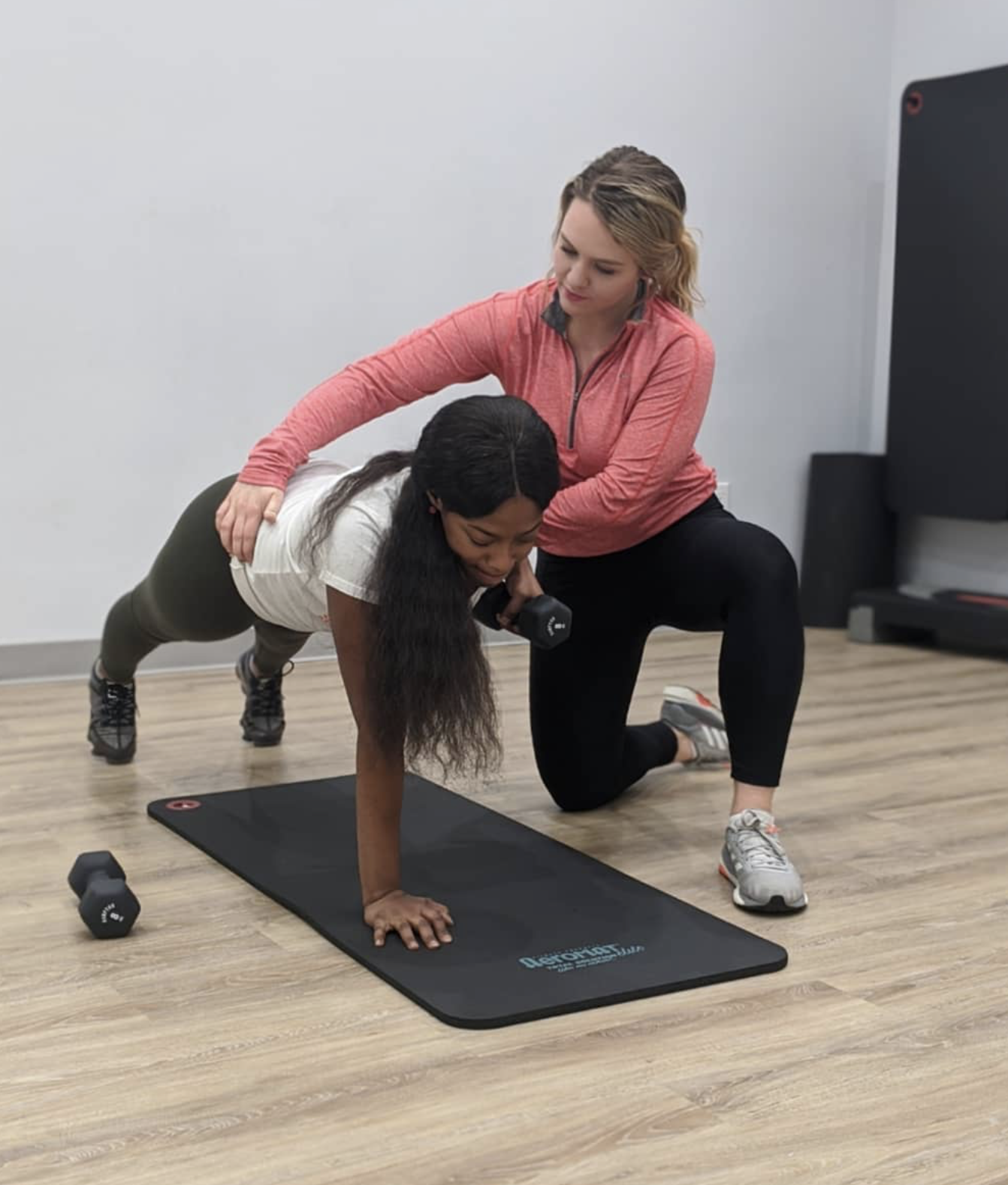 A woman assisting a woman performing a plank exercise with a dumbbell in a fitness studio.