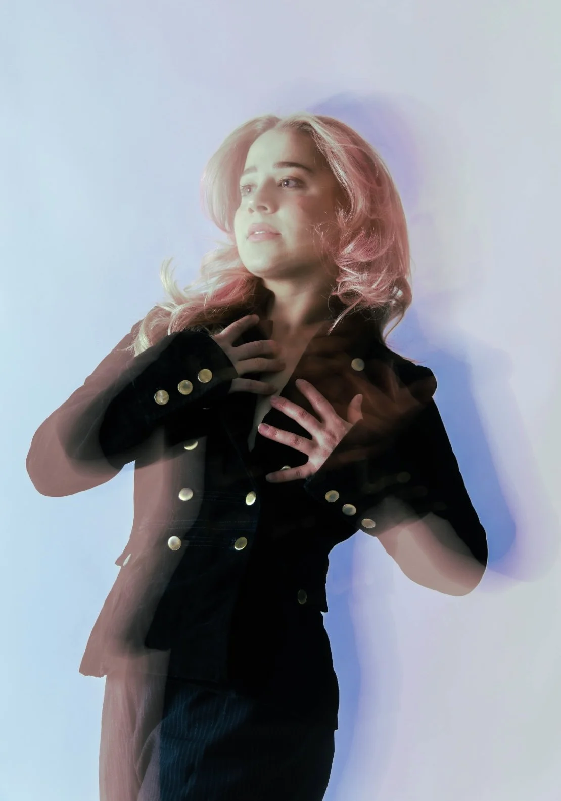 A woman with pink hair wearing a black jacket with gold buttons, posing with her hands on her chest, looking to the side against a plain background.