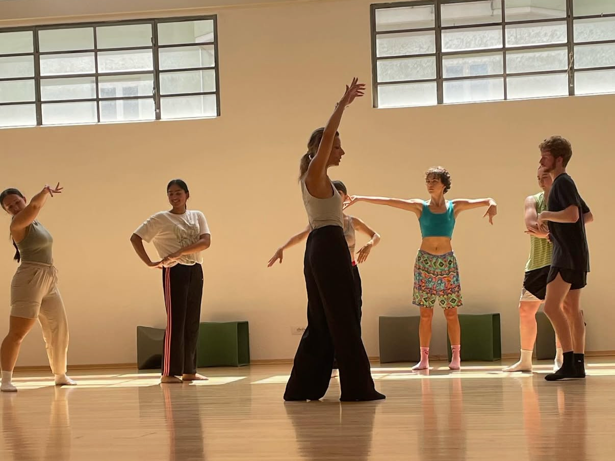 Group of six dancers practicing in a dance studio with wooden floors and large windows, some standing and some in dance poses.