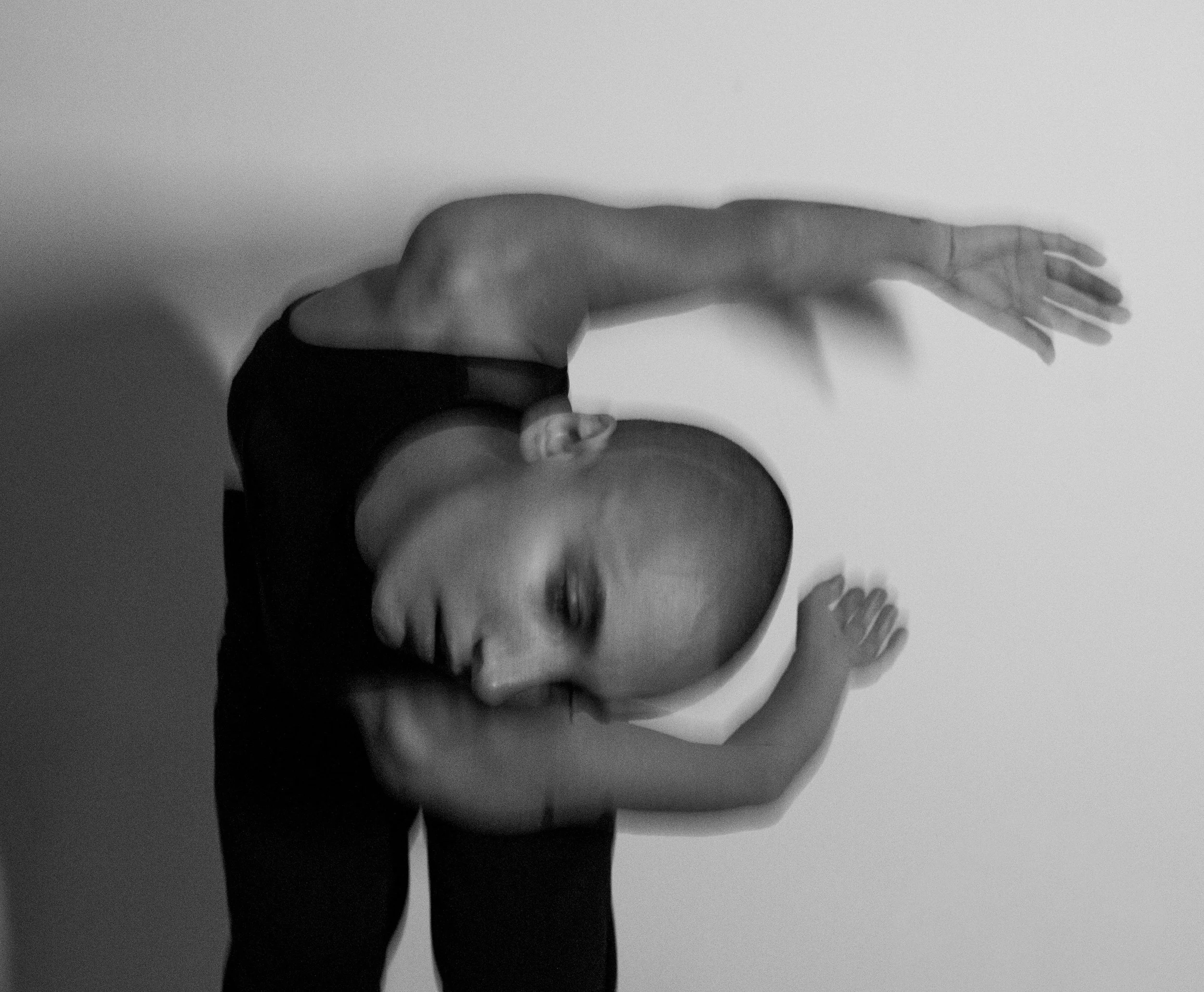 A person in a black top and pants is holding a baby upside down, supporting its back with one arm and touching its head with the other. The baby is facing downward with closed eyes, and the background is plain and white.