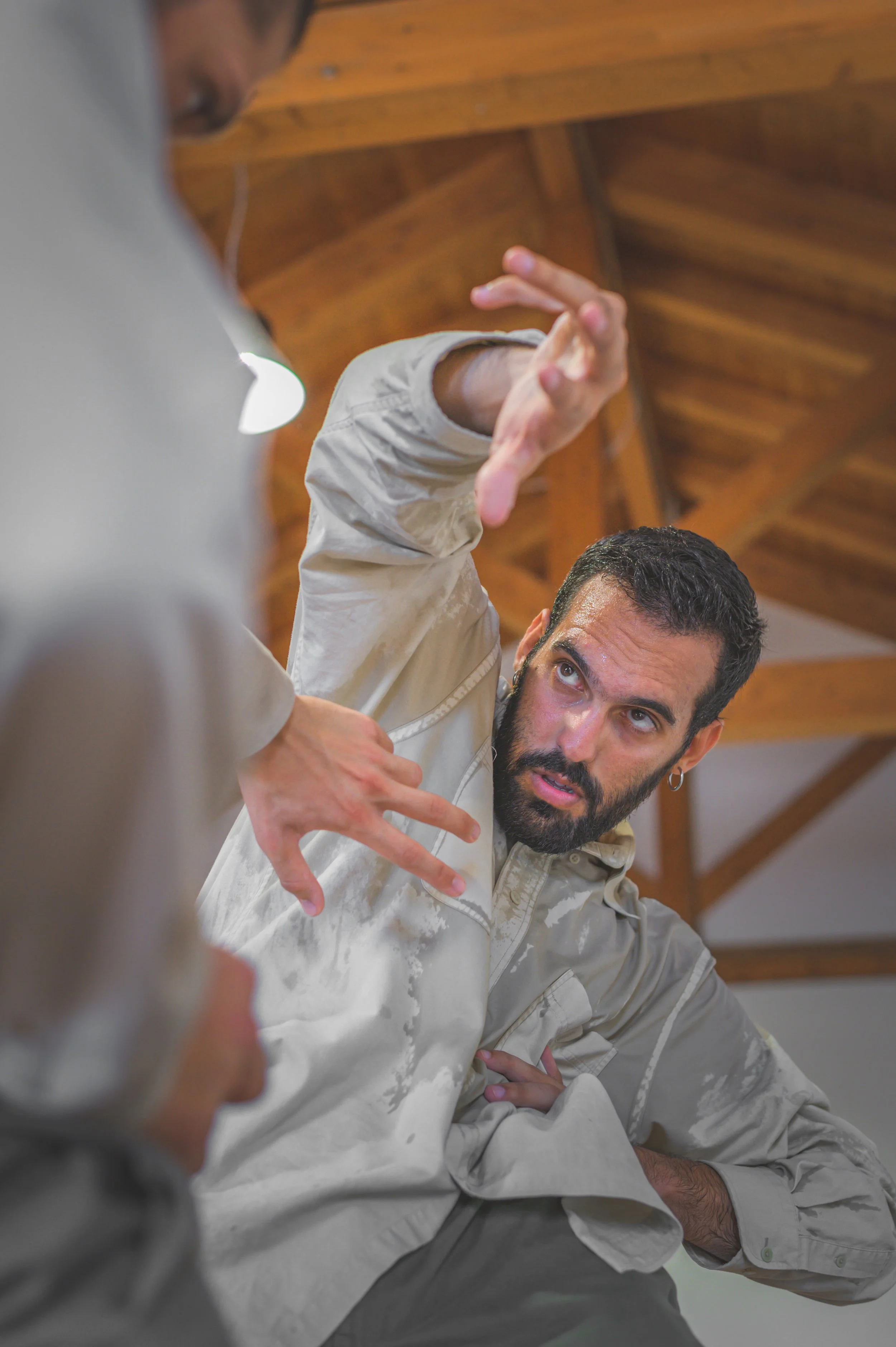 A man with a beard and earrings wearing a beige jacket is demonstrating a martial arts or self-defense move, blocking or controlling an opponent whose arm and hand are partially visible in the foreground. They are indoors with a wooden ceiling.