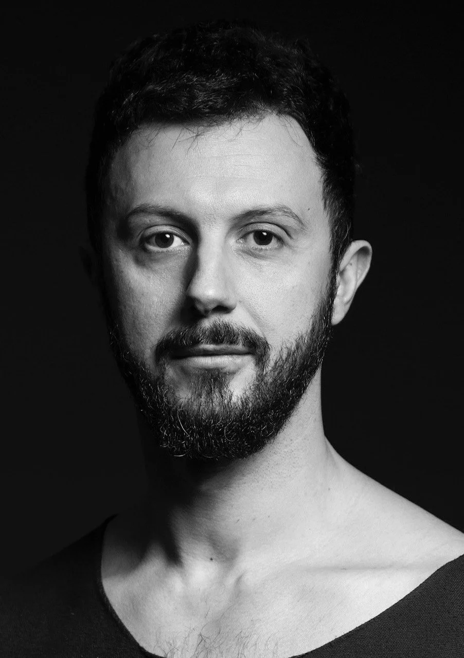 Black and white portrait of a man with short, dark hair and a beard, wearing a dark top, looking directly at the camera against a dark background.