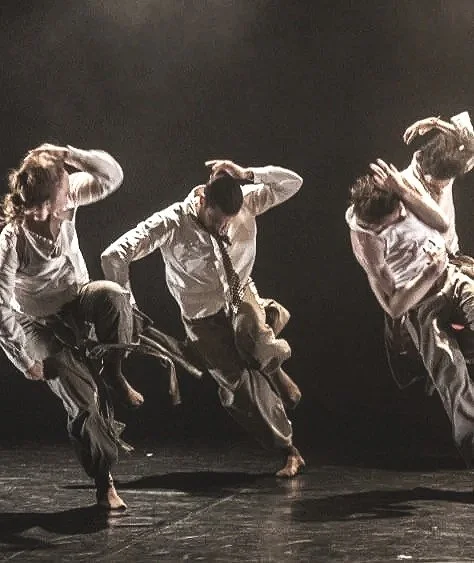Four dancers performing a contemporary dance on stage, each holding a puppet, with dramatic lighting and dark background.
