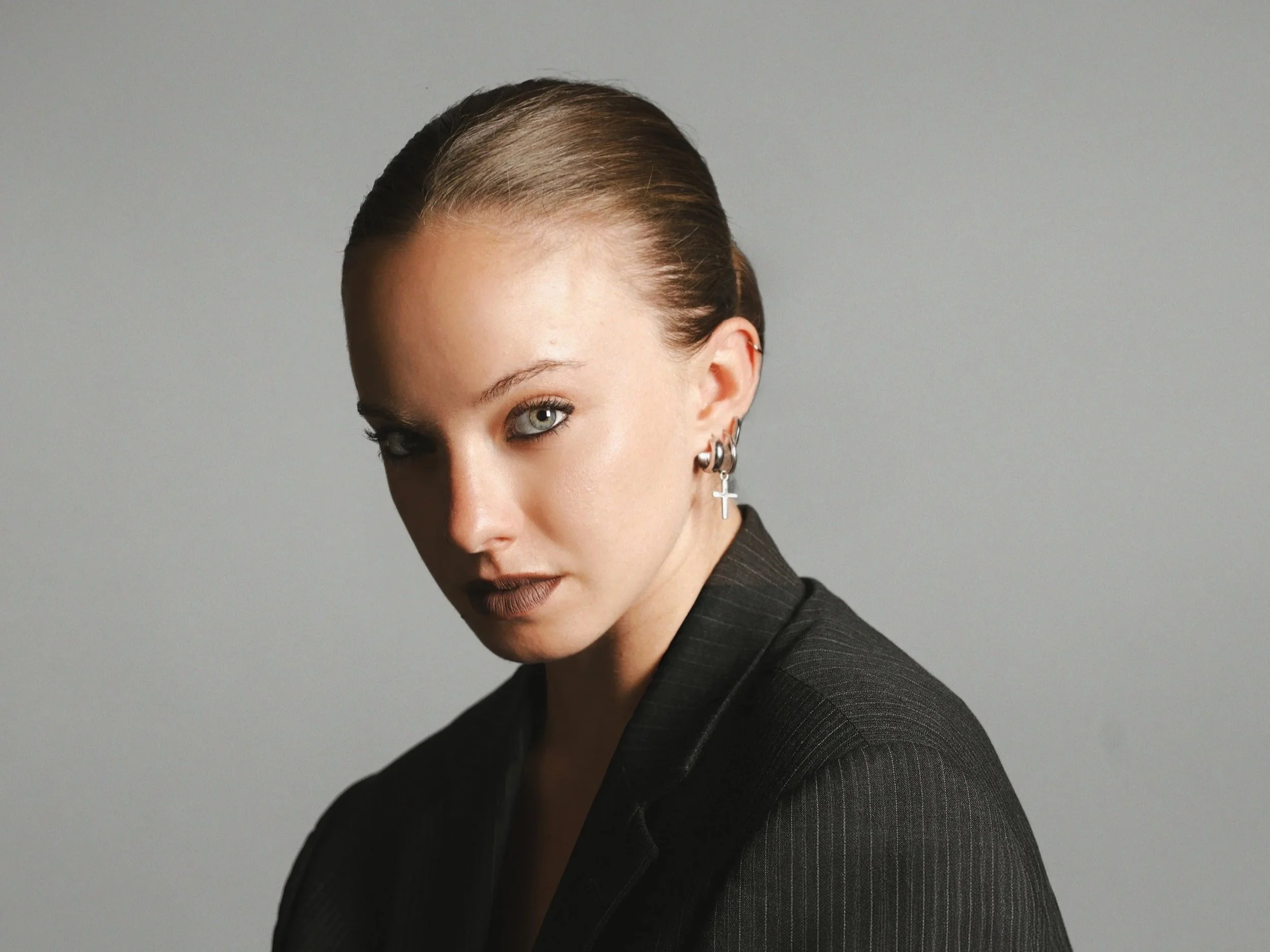 A woman with slicked-back hair wearing a black suit and multiple earrings, including a cross-shaped earring, posing against a grey background.